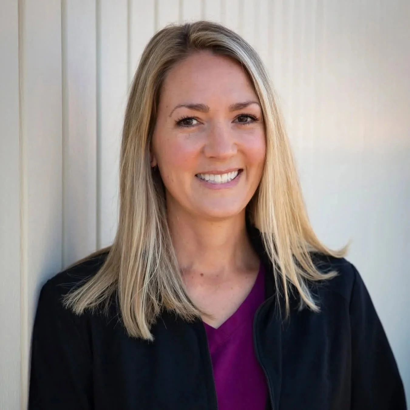 A woman with blonde hair smiling, wearing a black jacket over a purple top, standing against a light-colored wall.