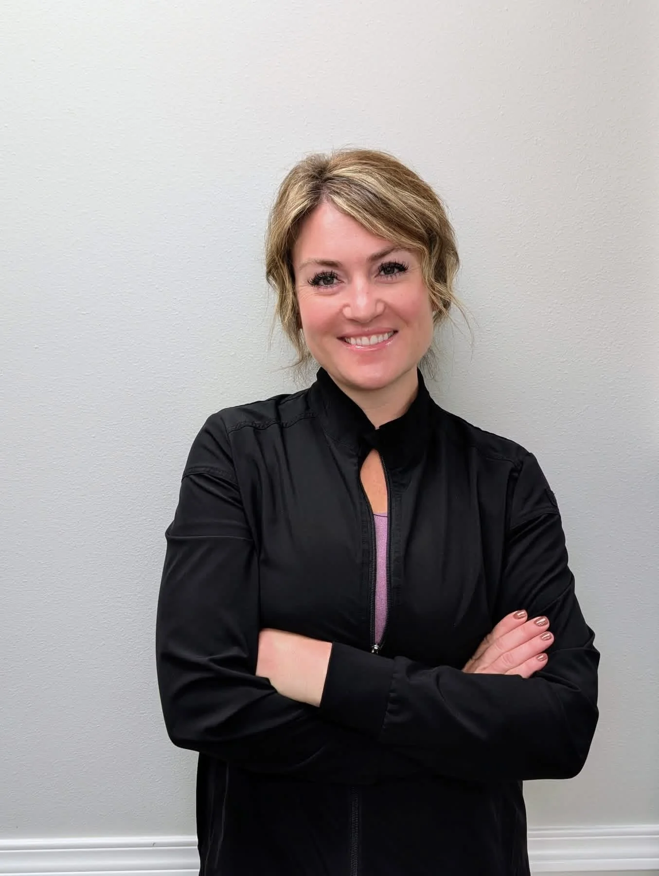 A woman with short wavy blonde hair, wearing a black jacket and a pink shirt underneath, standing against a plain light-colored wall. She is smiling with her arms crossed.