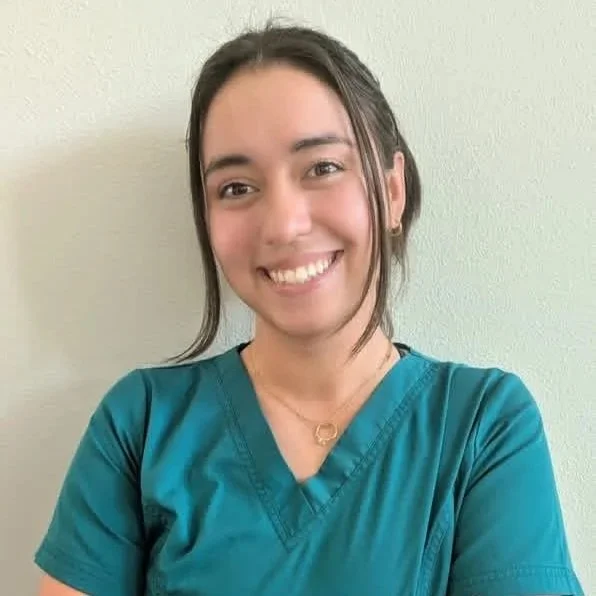A young woman wearing teal scrubs, smiling with her arms crossed, standing against a light-colored wall.