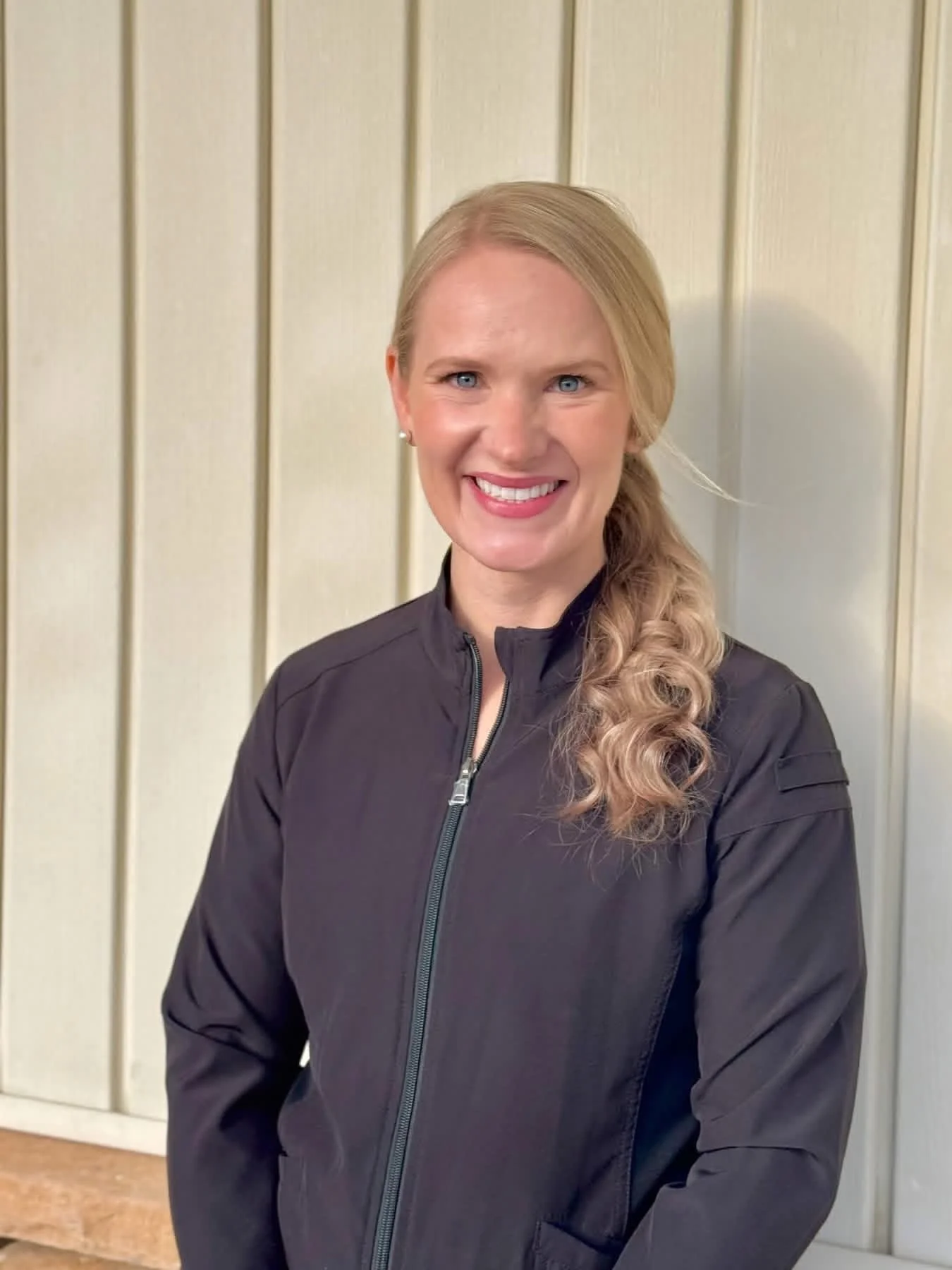 A woman with long blonde hair styled in a loose side braid, smiling, wearing a black zip-up jacket, standing in front of a cream-colored wood panel wall.