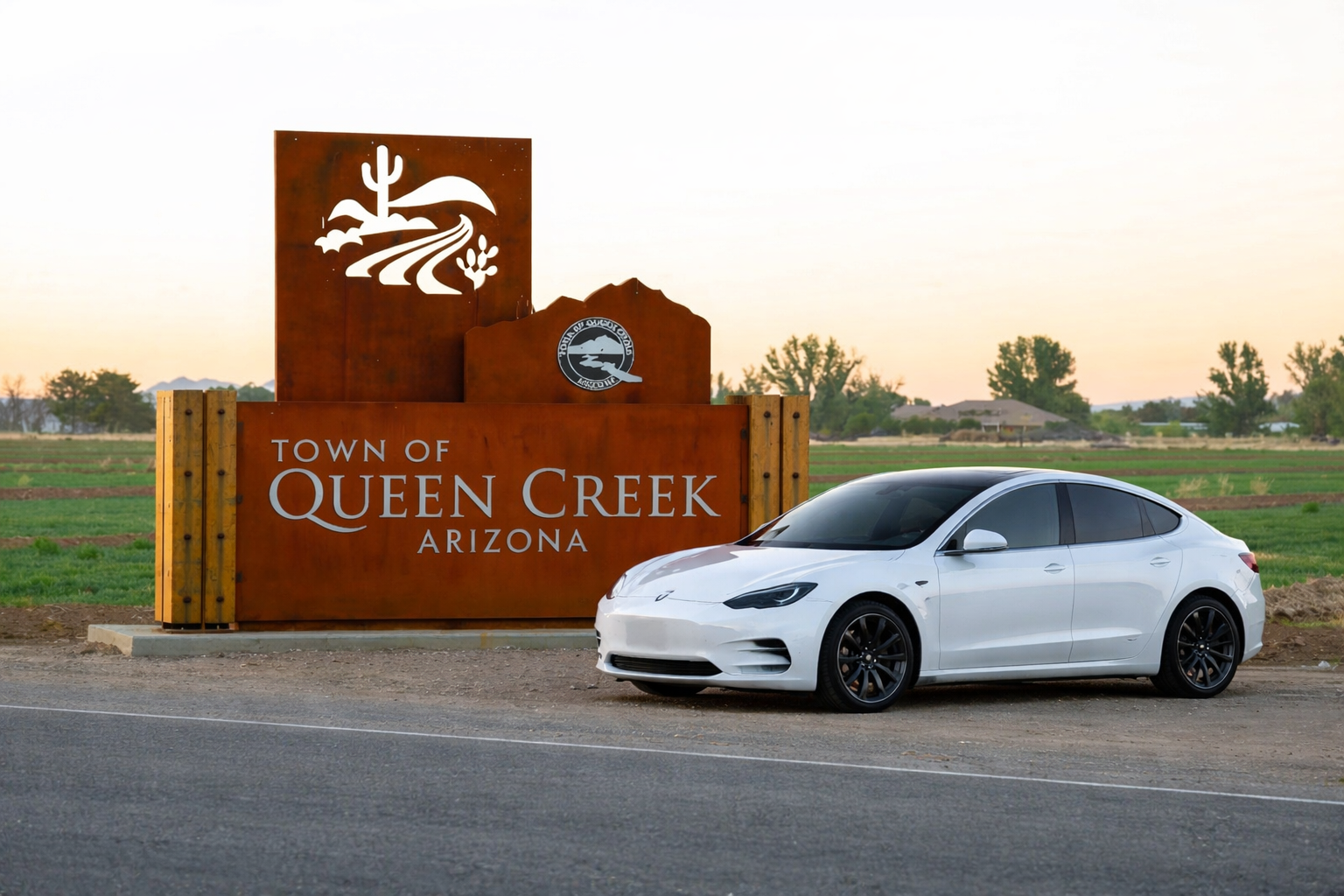 White sedan parked beside the sign for the town of Queen Creek, Arizona, with a backdrop of open fields and trees during sunset.