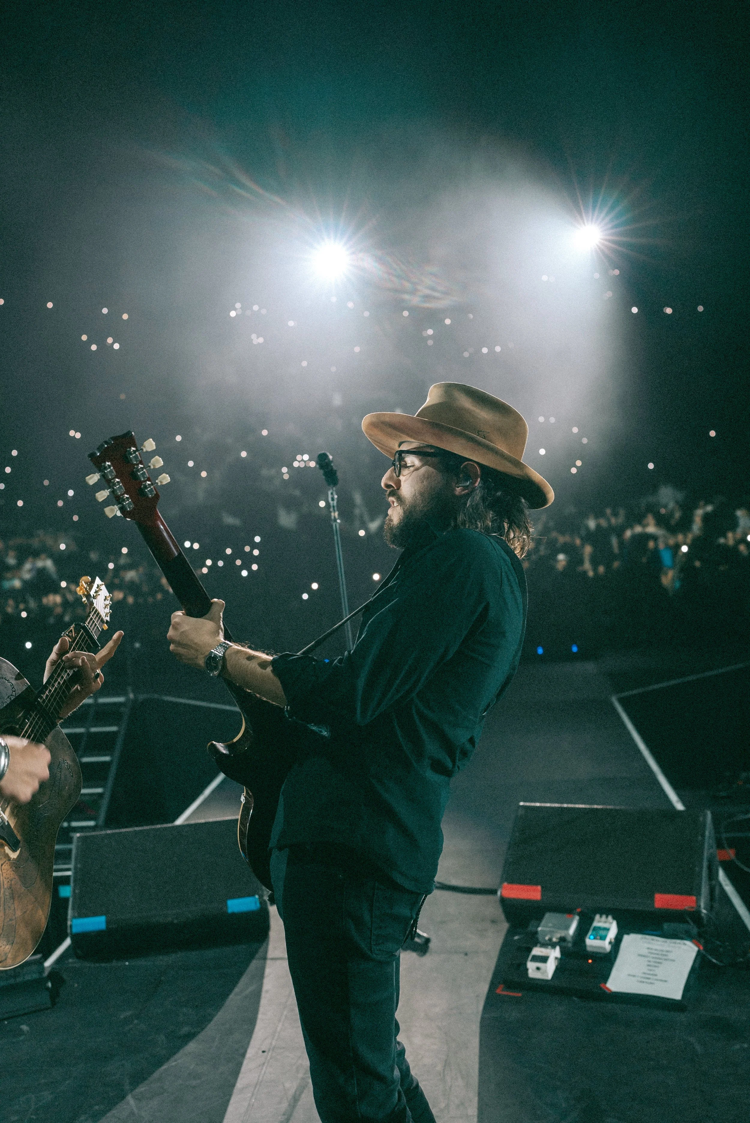 Musician with a beard, glasses, and a tan hat playing an electric guitar on stage with bright lights and an audience with cell phone lights in the background.