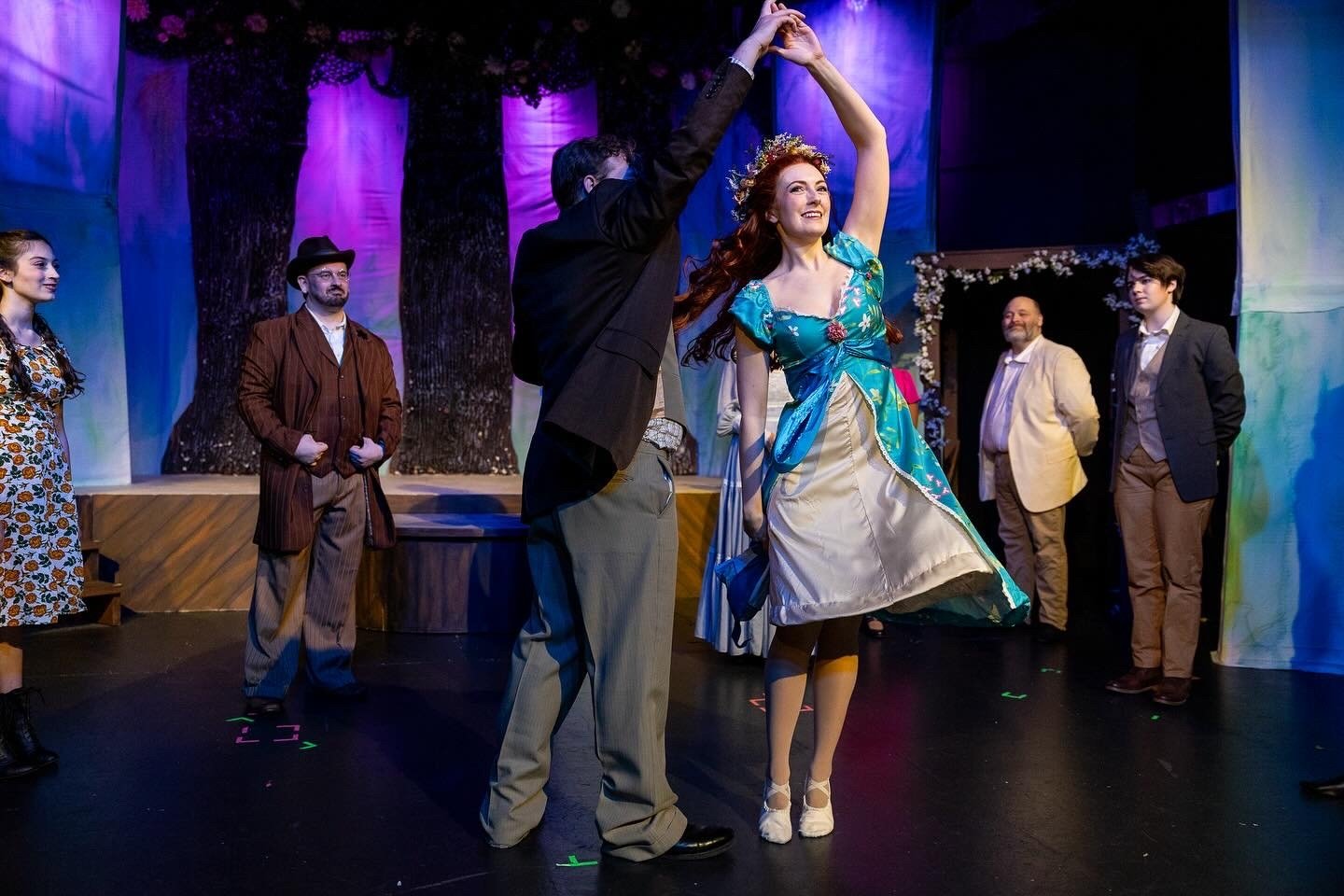 A theatrical stage scene with a man and woman dancing, surrounded by other seated and standing actors in vintage costumes. The woman wears a colorful dress with a floral crown, and the man is in dark formal attire. The stage background is decorated w