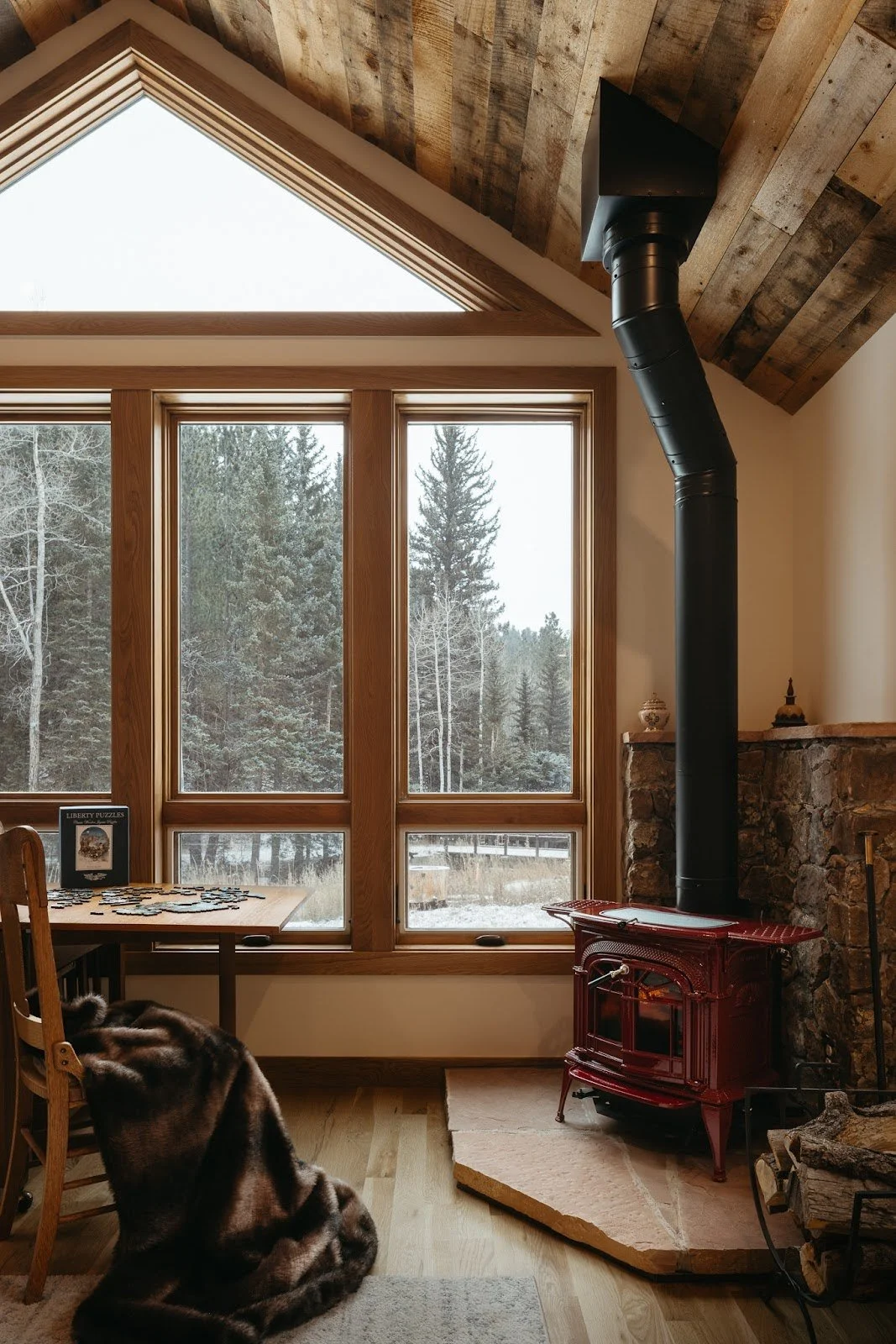 Cozy cabin interior with large windows, wood-burning stove and forest views, showcasing warm natural materials and natural light