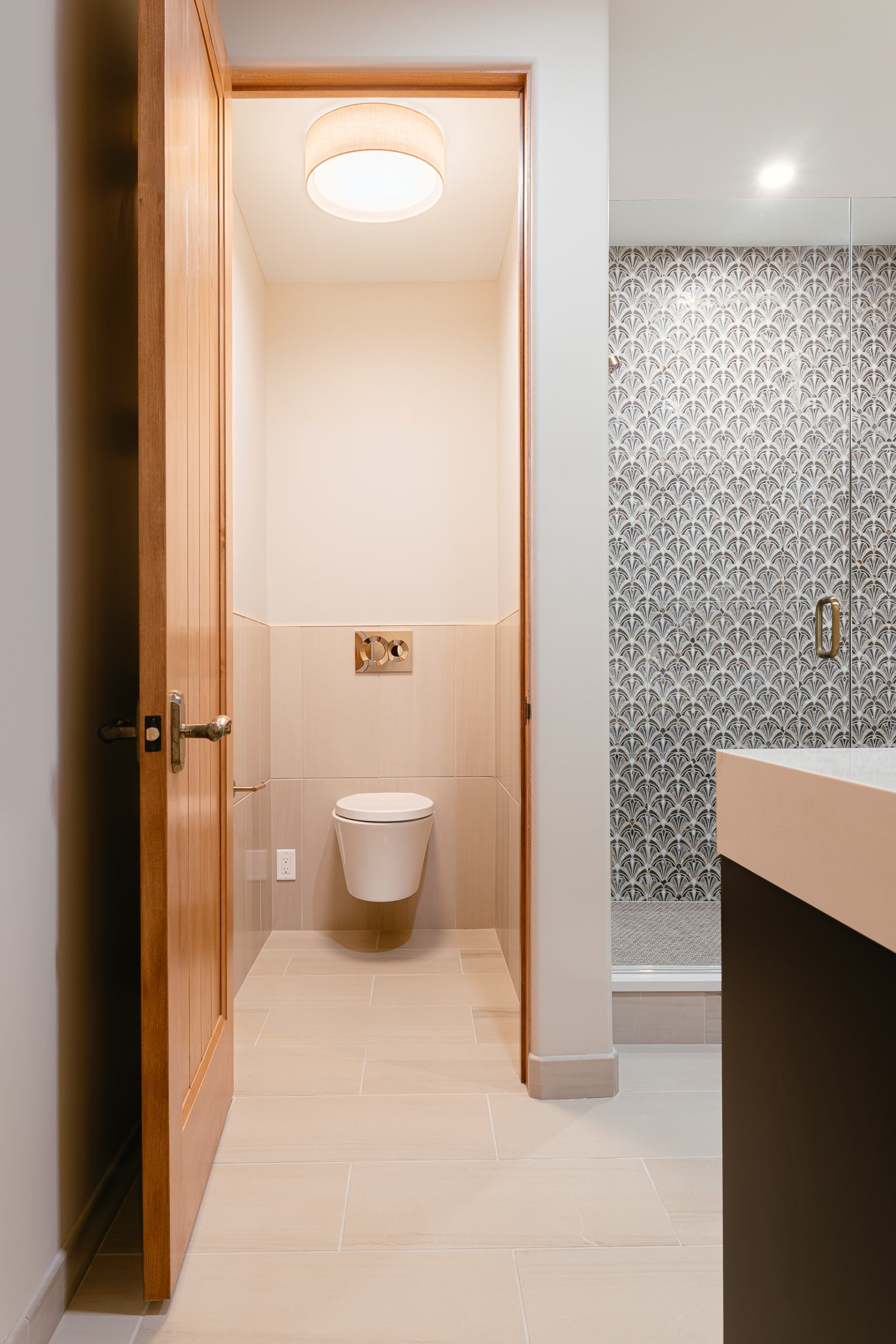 Modern bathroom with wooden door, wall-mounted toilet, patterned shower wall, and beige tile flooring.