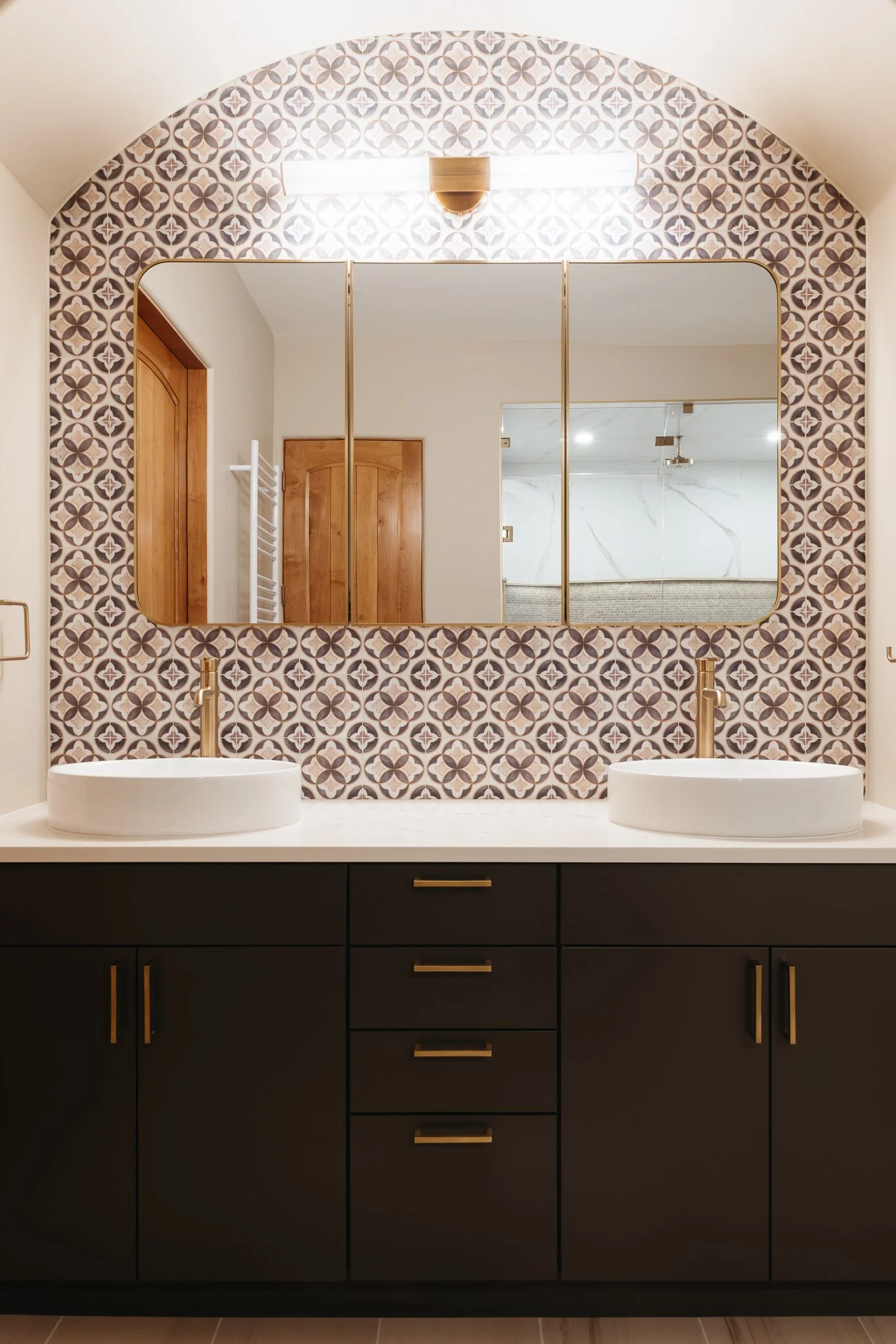 Modern bathroom with dark vanity, double white sinks, patterned tile backsplash, and large mirrors.