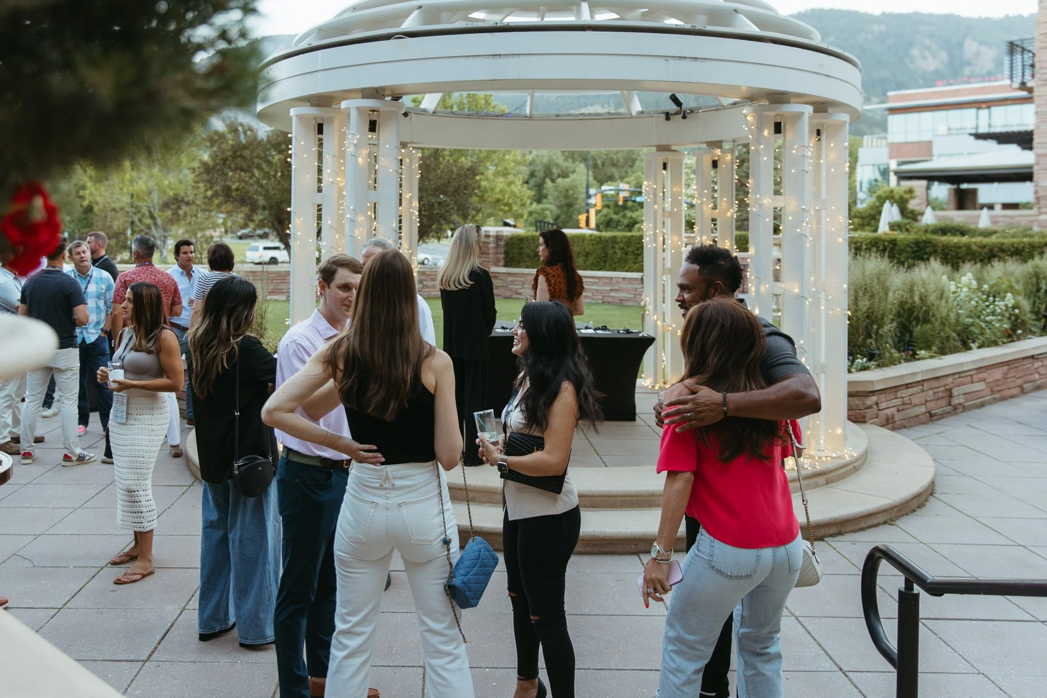 Group of people socializing outdoors at an event under a gazebo with string lights.