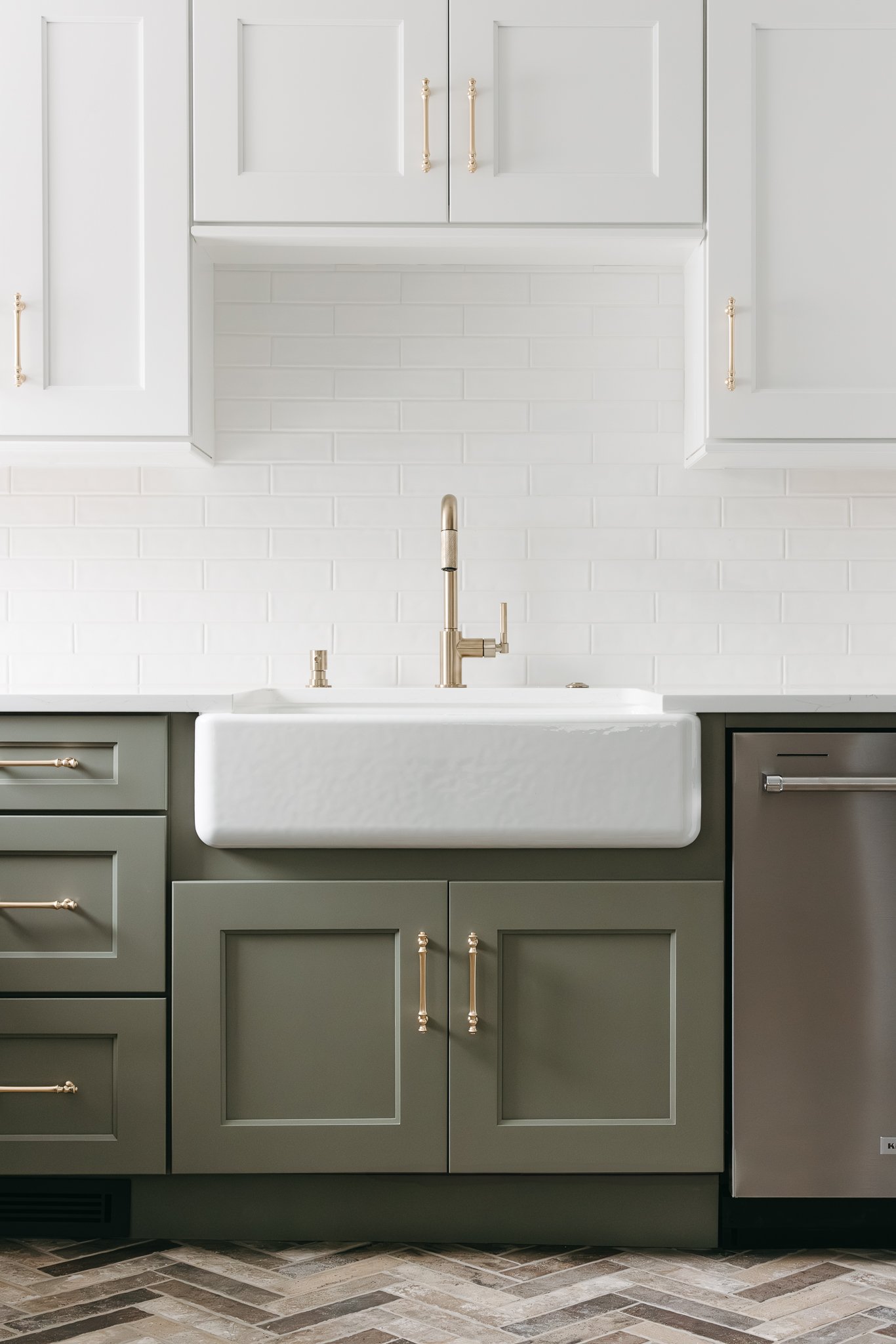 Modern kitchen with a white farmhouse sink, gray cabinets, brass hardware, subway tile backsplash, and herringbone floor.