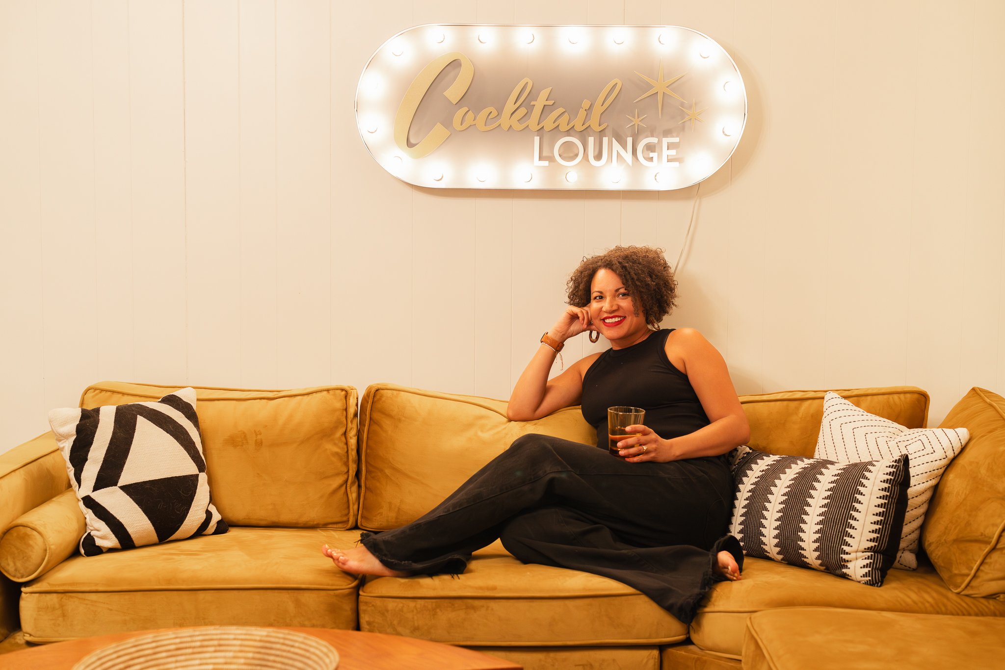 woman wearing all black sitting on a vintage mustard yellow sofa, relaxed with one hand rested on the side of her face and the other holding a cocktail with a lit up "cocktail lounge" sign hanging above her on the white-painted wood panel wall