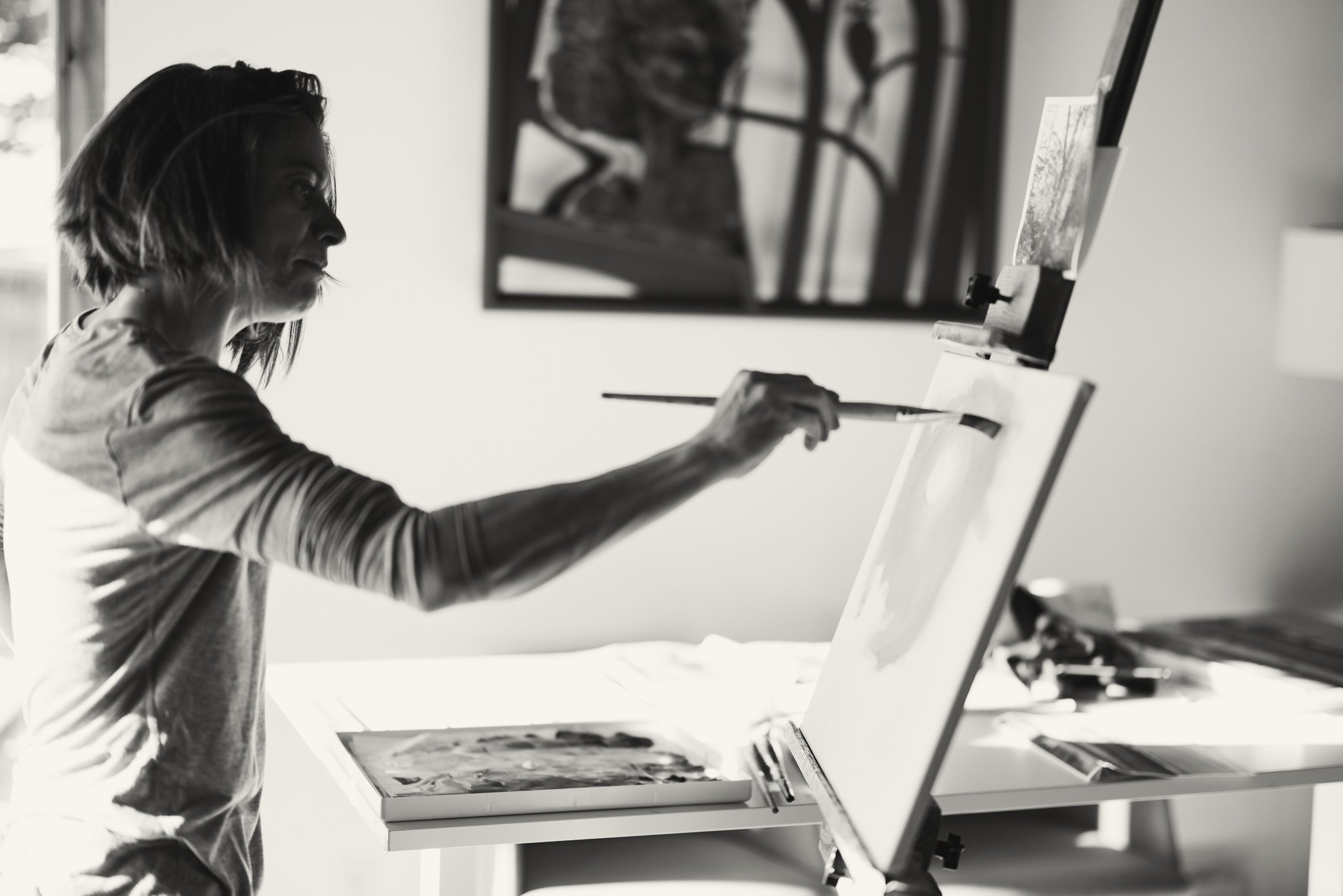 Artist painting on a canvas in a studio, using a brush, with a palette on the table. Black and white photo.