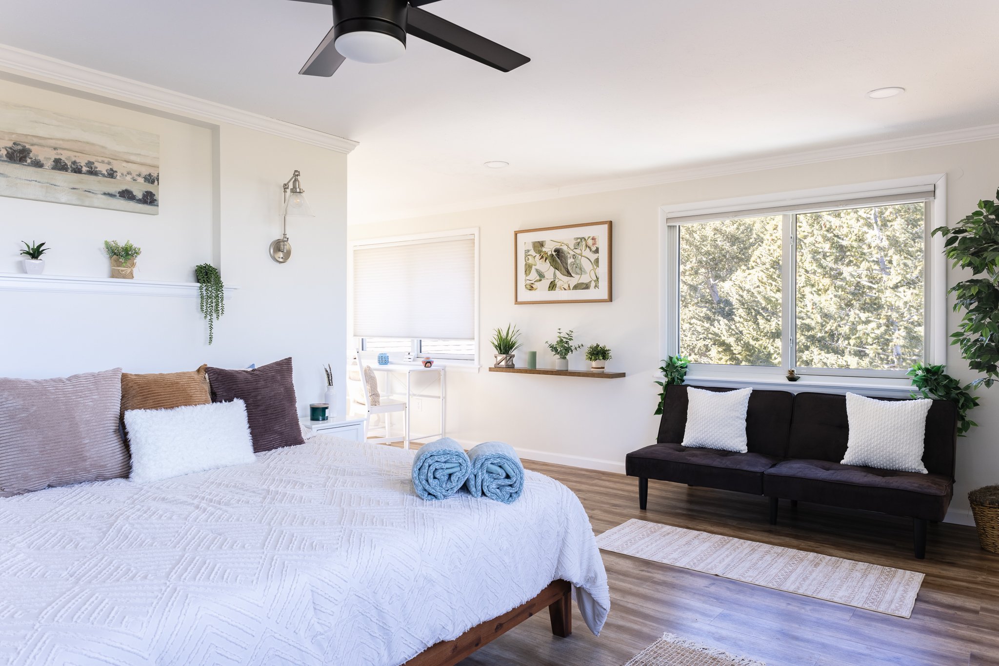 Bright, modern bedroom with a bed featuring brown and white pillows, a white comforter, and two rolled blue towels. There's a sofa with white cushions, decorative plants, a wall shelf, framed artwork, a ceiling fan, and large windows with natural lig