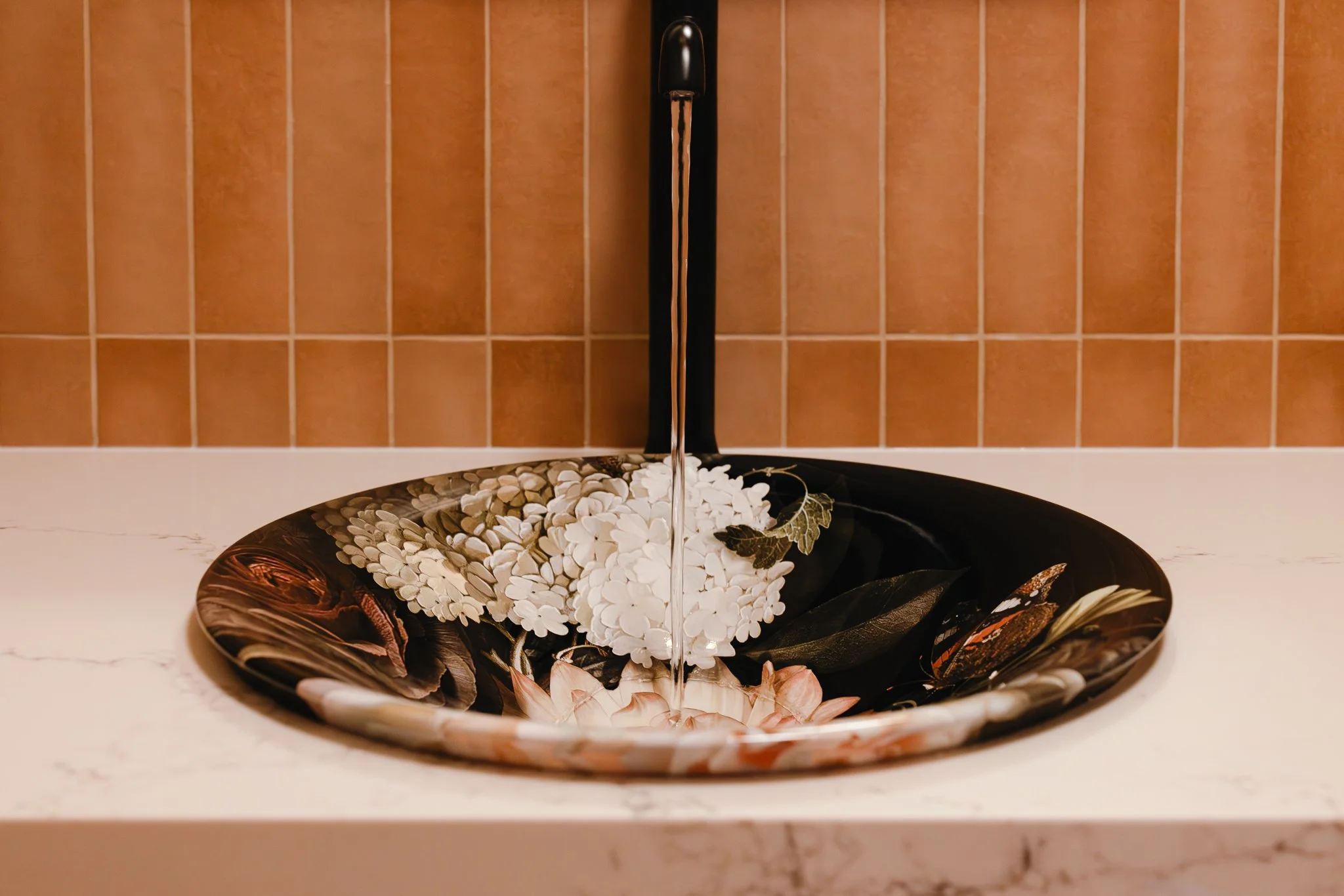 Decorative vessel sink with floral design and black faucet against warm terracotta tile backsplash.