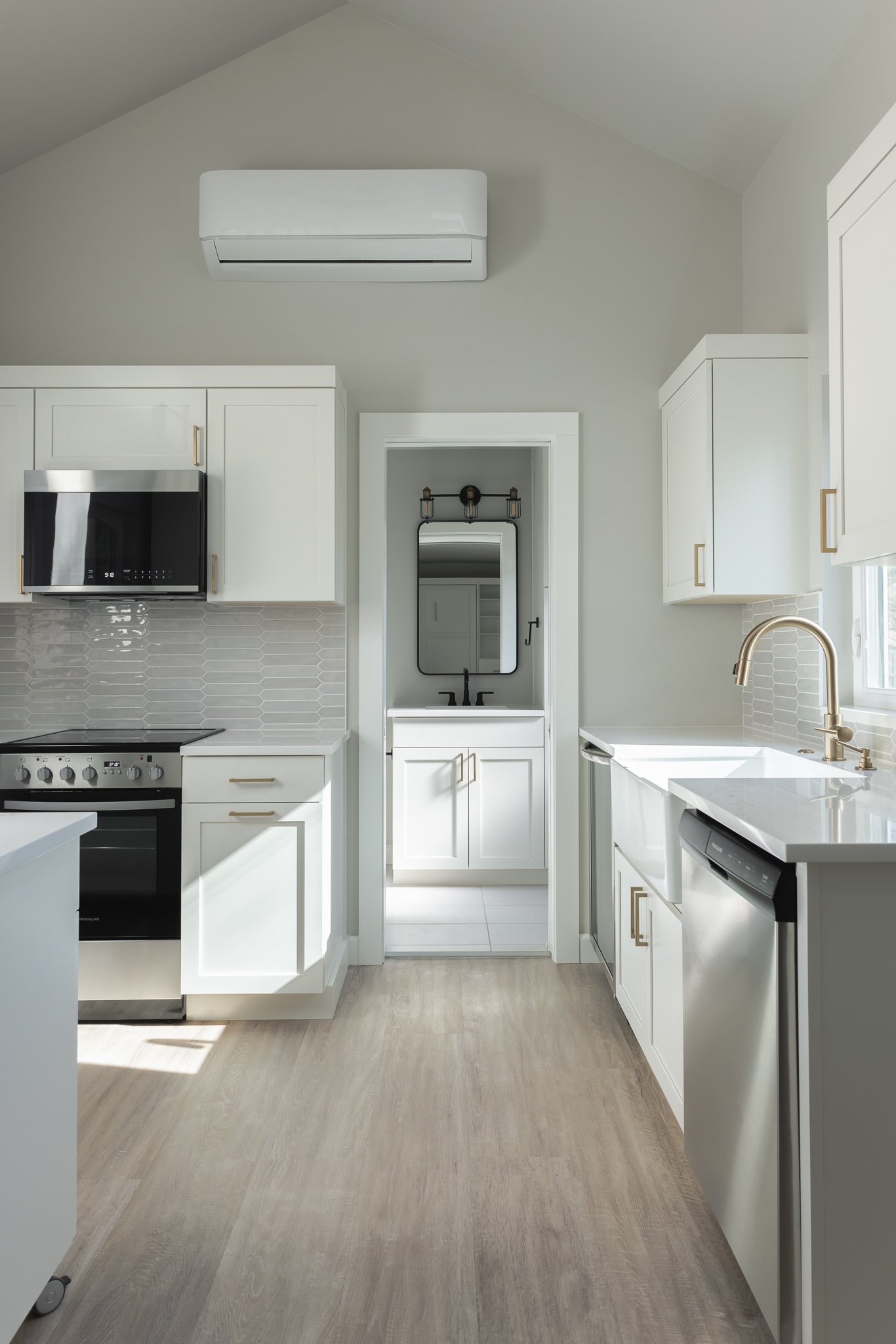 Modern kitchen interior with white cabinets, stainless steel appliances, a dishwasher, and a gray backsplash. There's a doorway leading to a bathroom with a mirror and vanity. The floor has a light wood finish, and an air conditioning unit is mounted