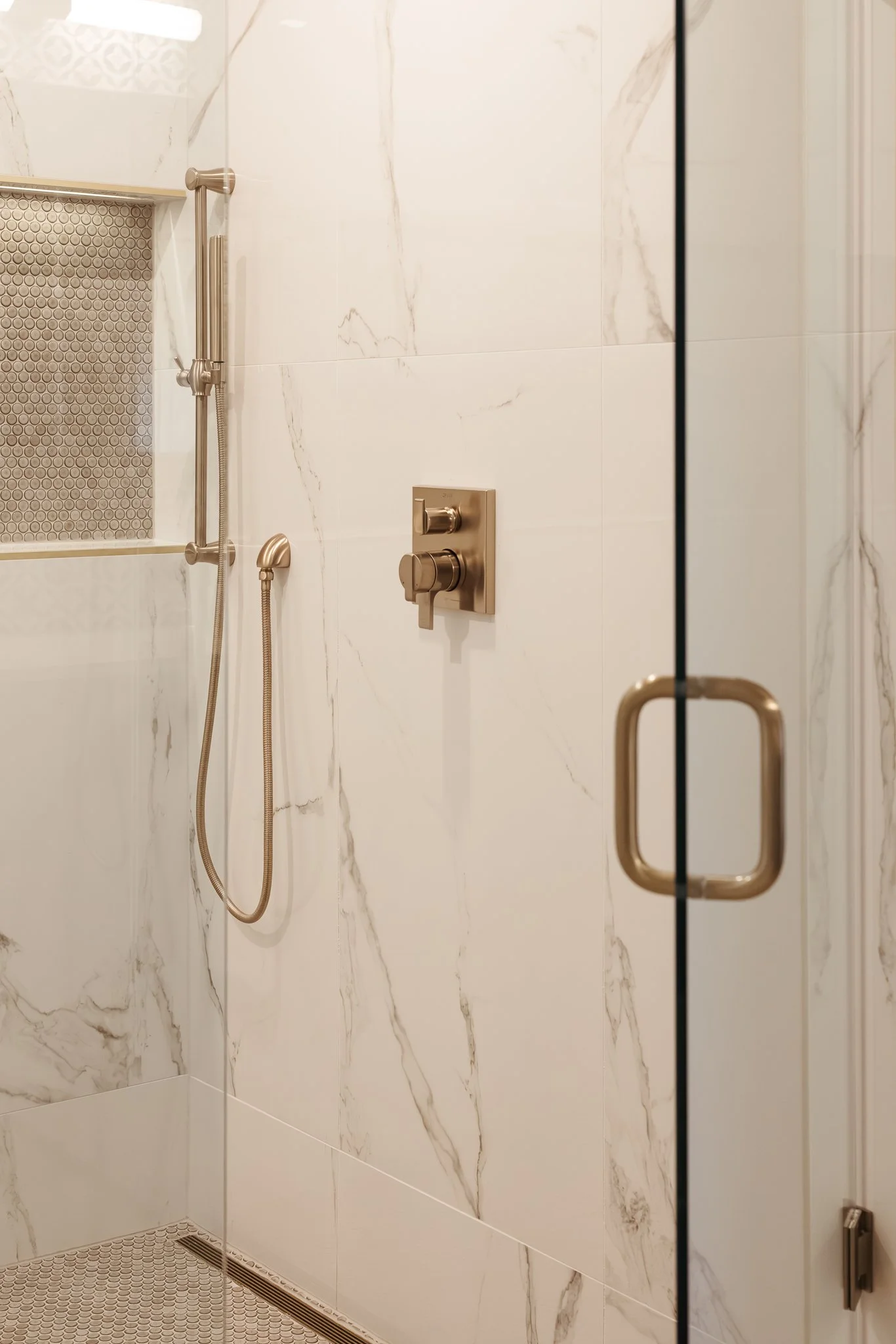 Modern bathroom shower with marble wall tiles, brass shower head and controls, and glass door.