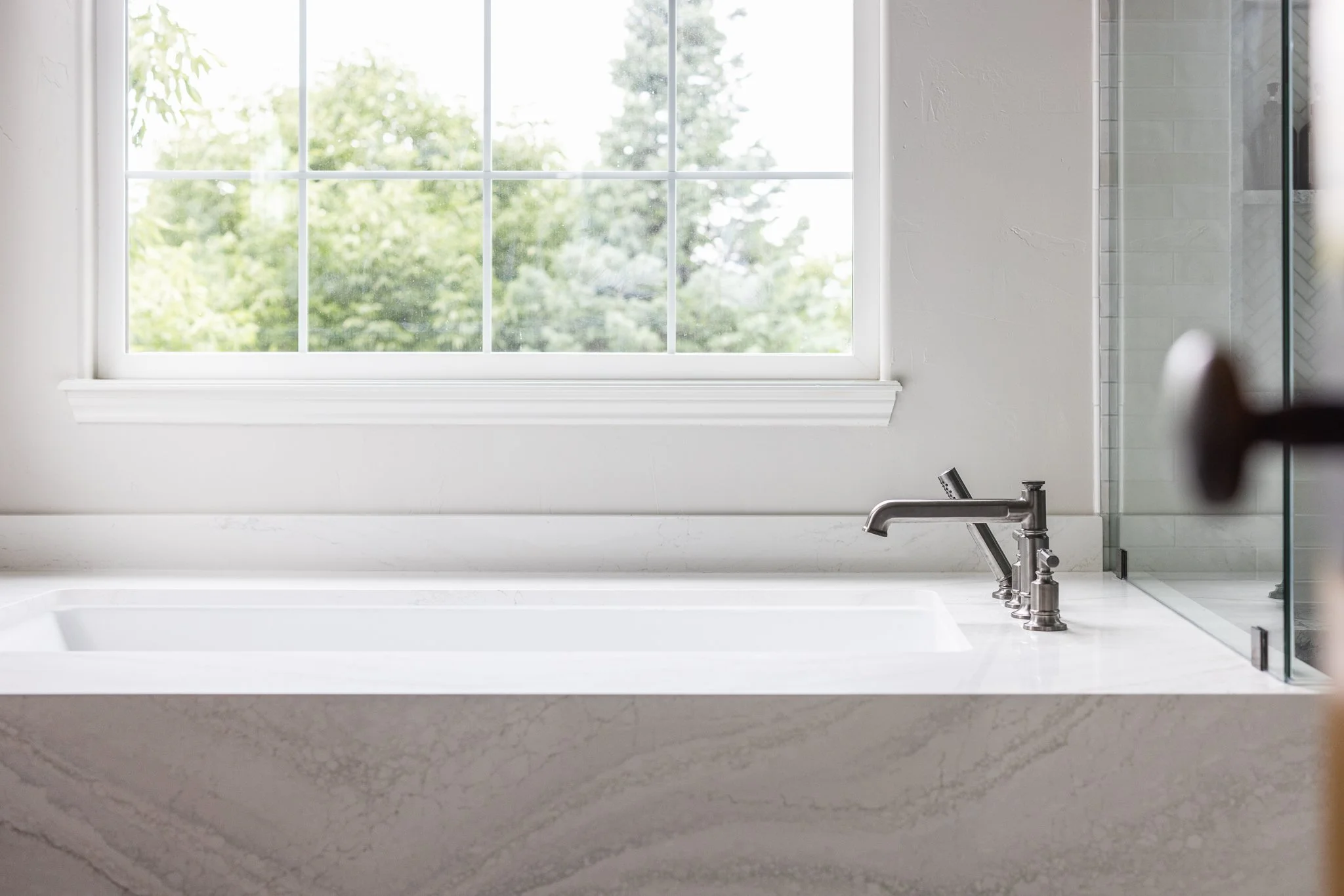 White soaking tub with marble surround and chrome faucet beneath a large window, photographed in a bright residential bathroom.