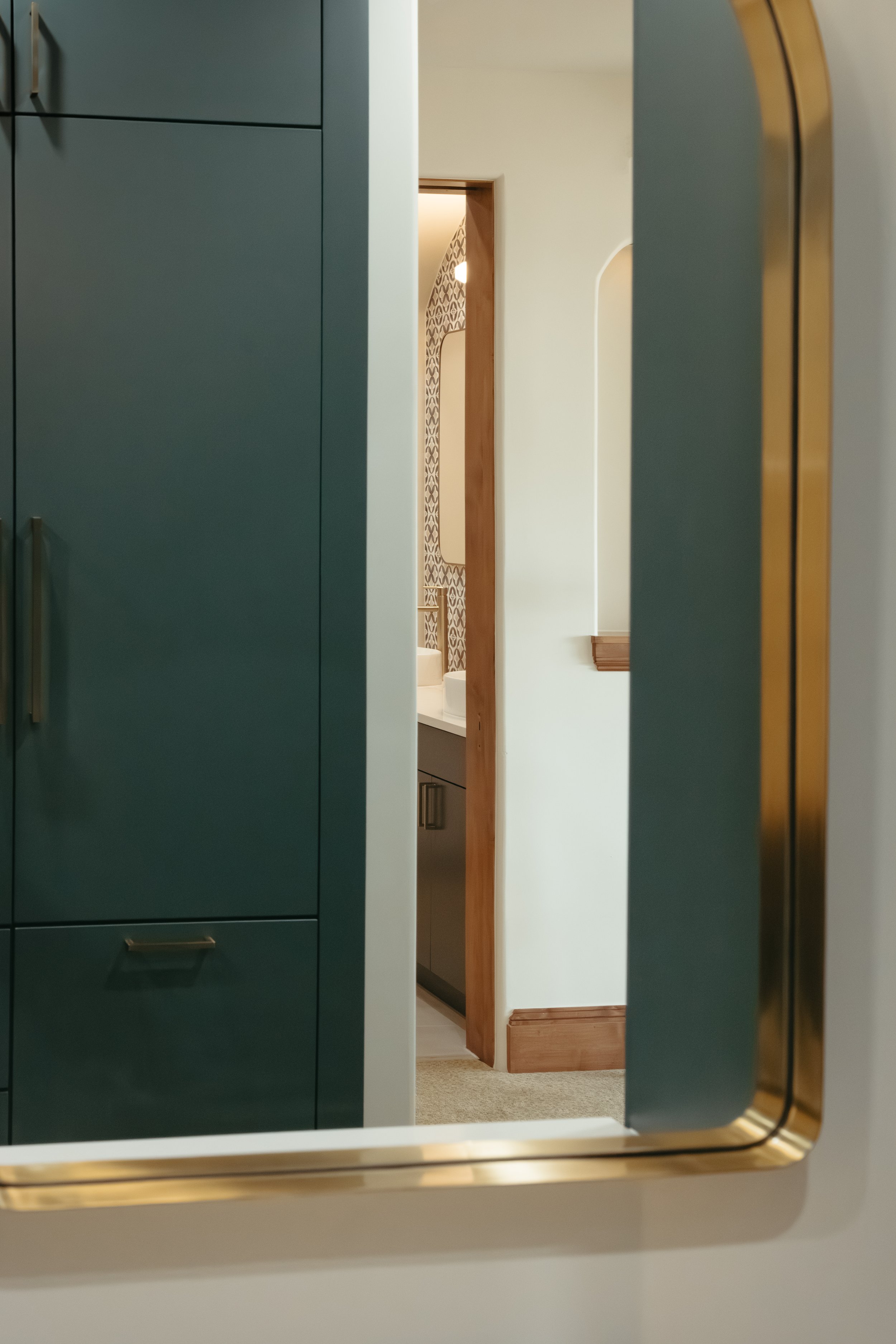 Interior view through a doorway showing a modern bathroom with a sink and patterned wall tiles, including a dark green cabinet and a gold-framed mirror.