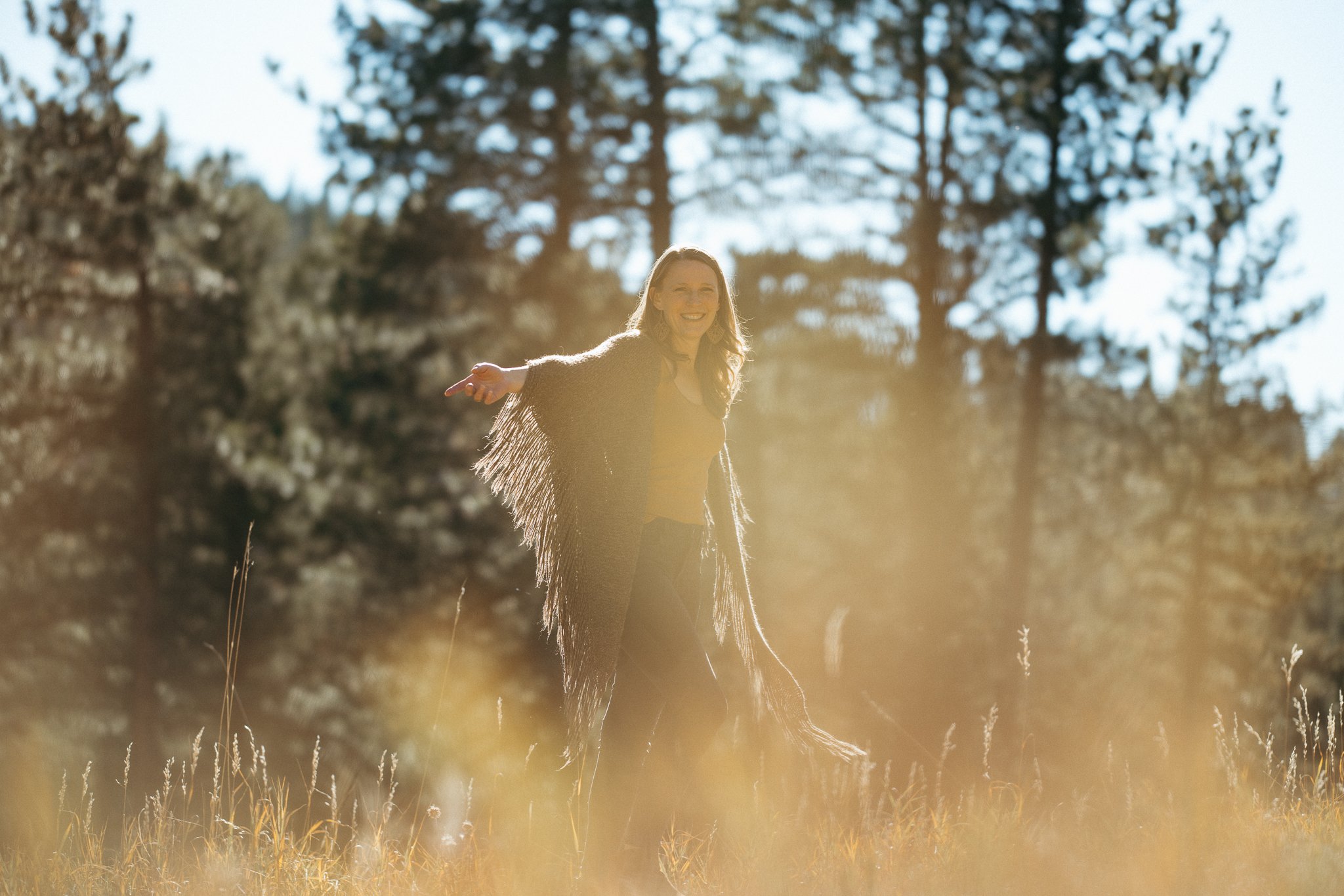 Person in a field with trees in the background, wearing a fringed shawl, smiling in sunlight.