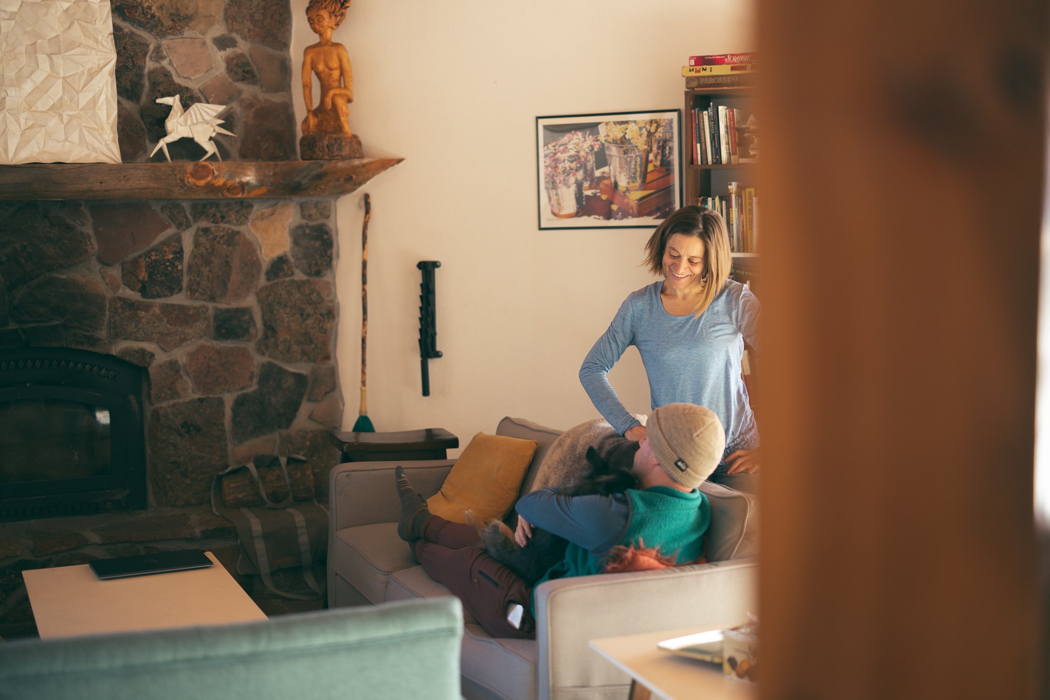 A cozy living room scene with a stone fireplace, wooden mantel, and decorative items. A person is lying on a couch with another person standing nearby, smiling. Books and framed pictures are on the wall.