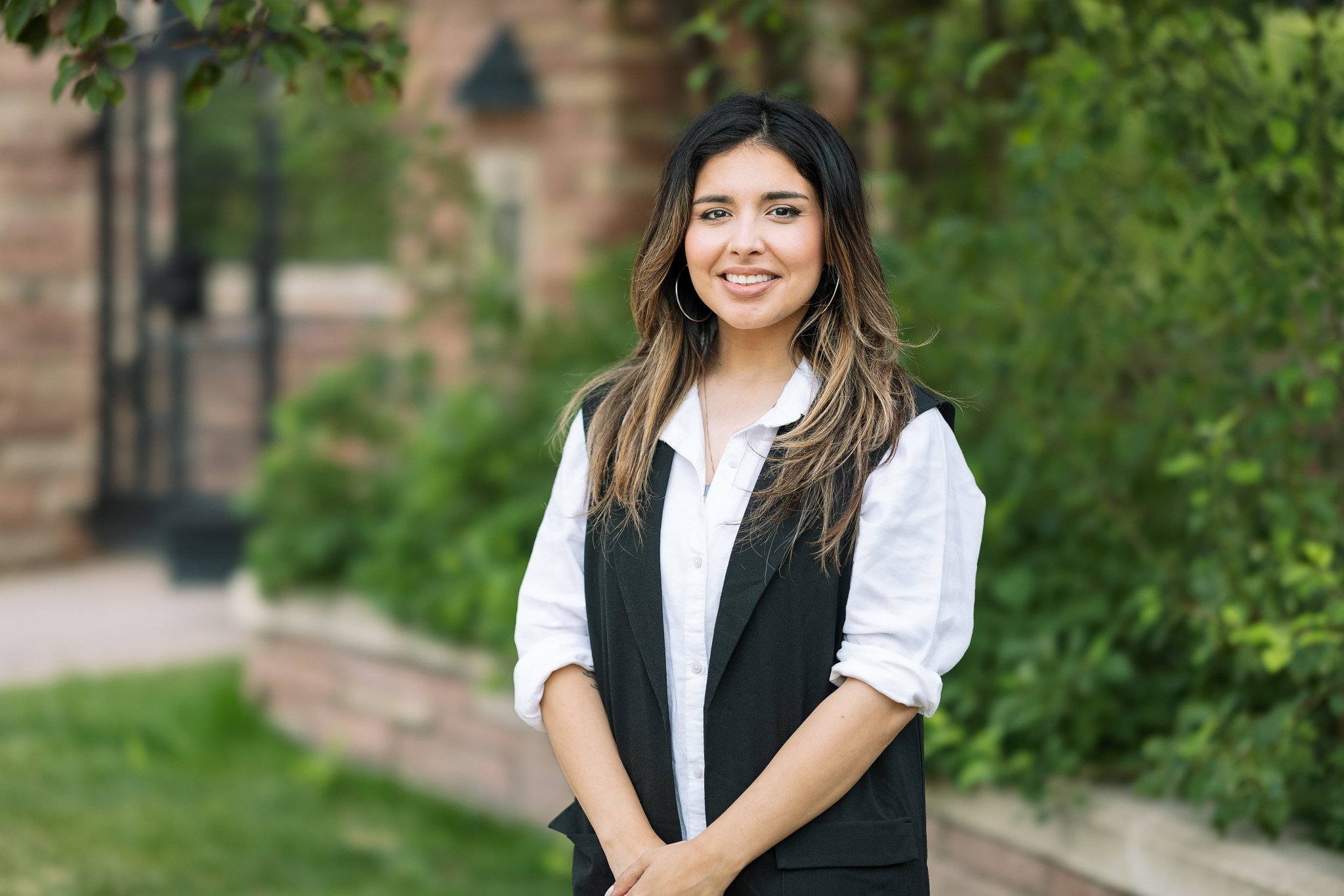 Smiling person standing outdoors with green foliage in the background, wearing a black vest and white shirt.