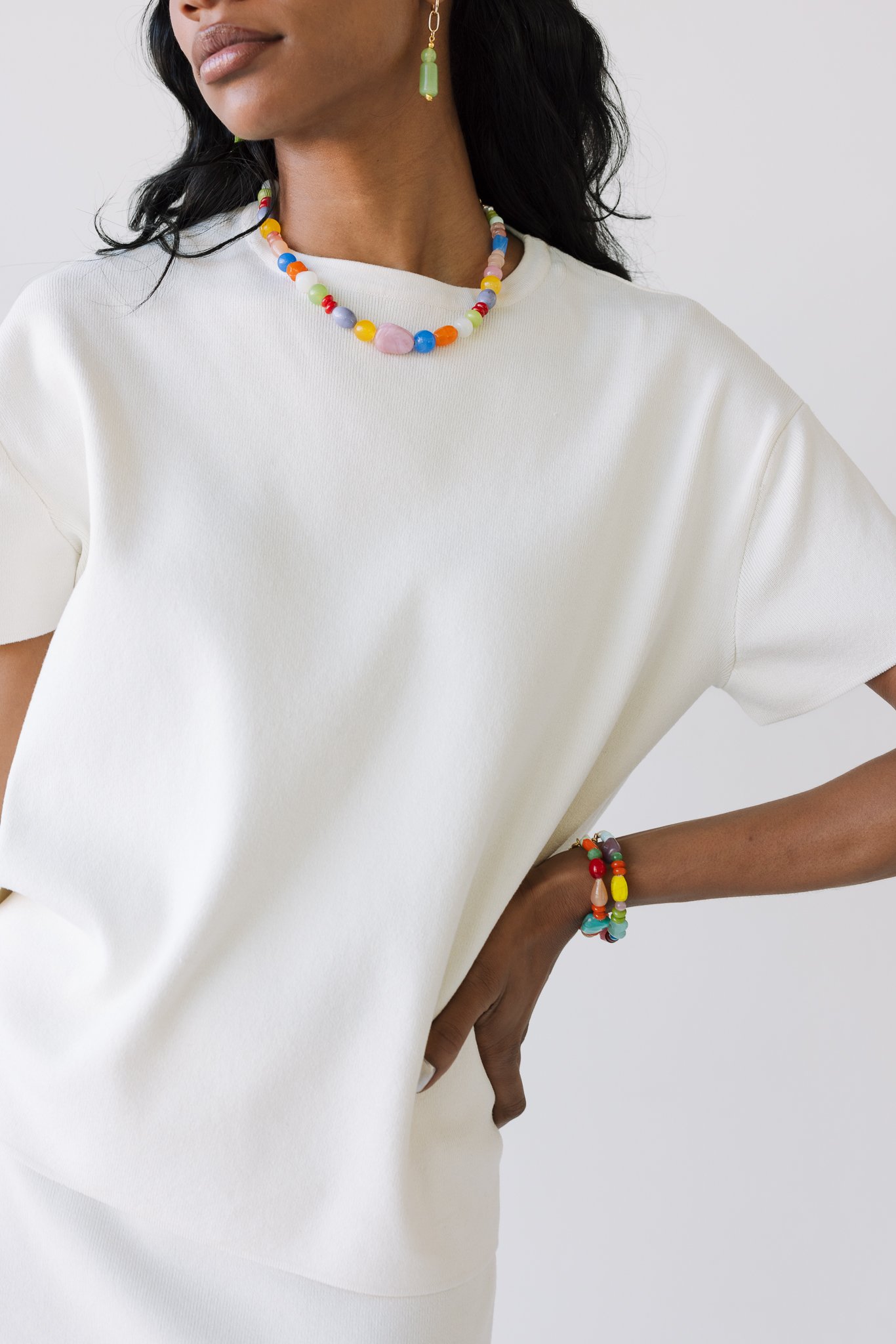 Person wearing a white t-shirt with a colorful beaded necklace, multi-colored beaded bracelet, and a single green dangling earring.