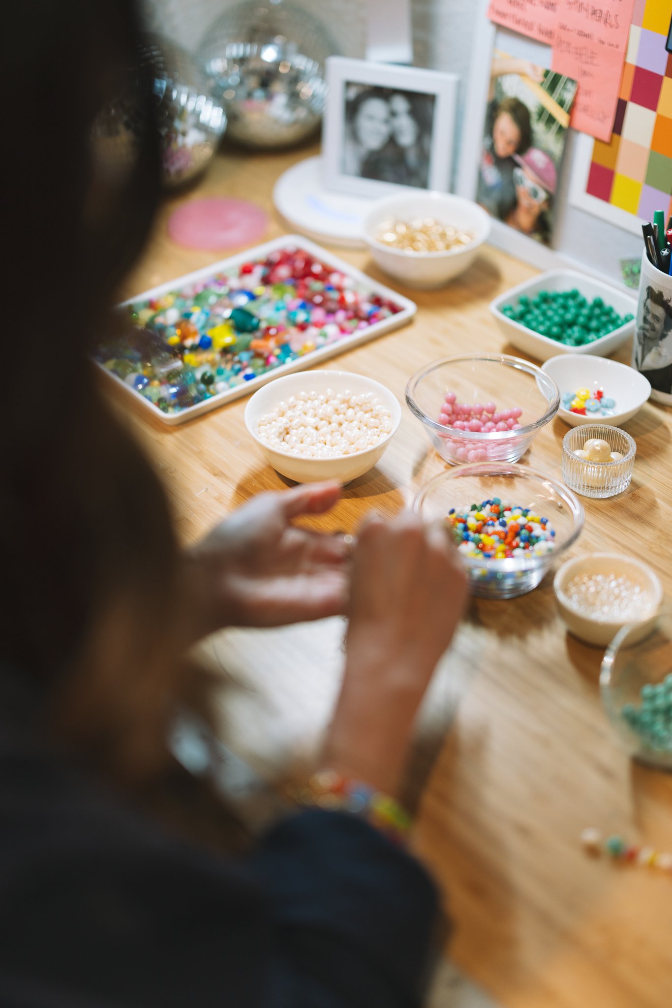 A person arranging colorful beads on a wooden table, with bowls containing various beads. There are photographs and decorative items in the background.
