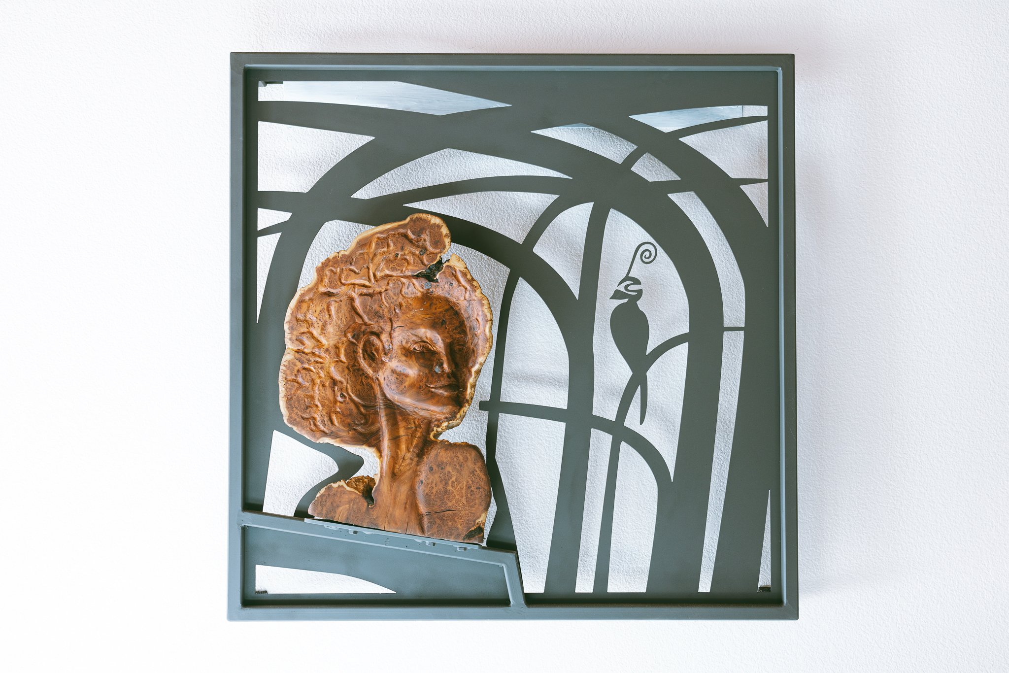 Art sculpture featuring a carved wooden female face with curly hair integrated into a black metal frame with abstract geometric cutouts and a stylized bird silhouette.