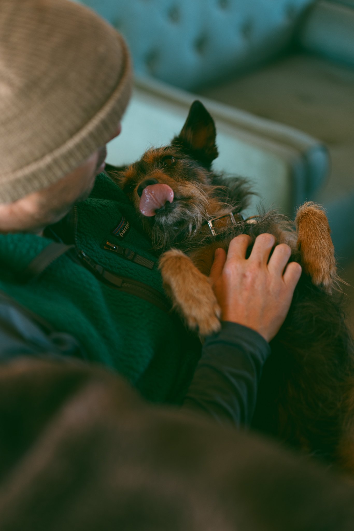 Person in a beanie holding a small dog that is licking its nose.