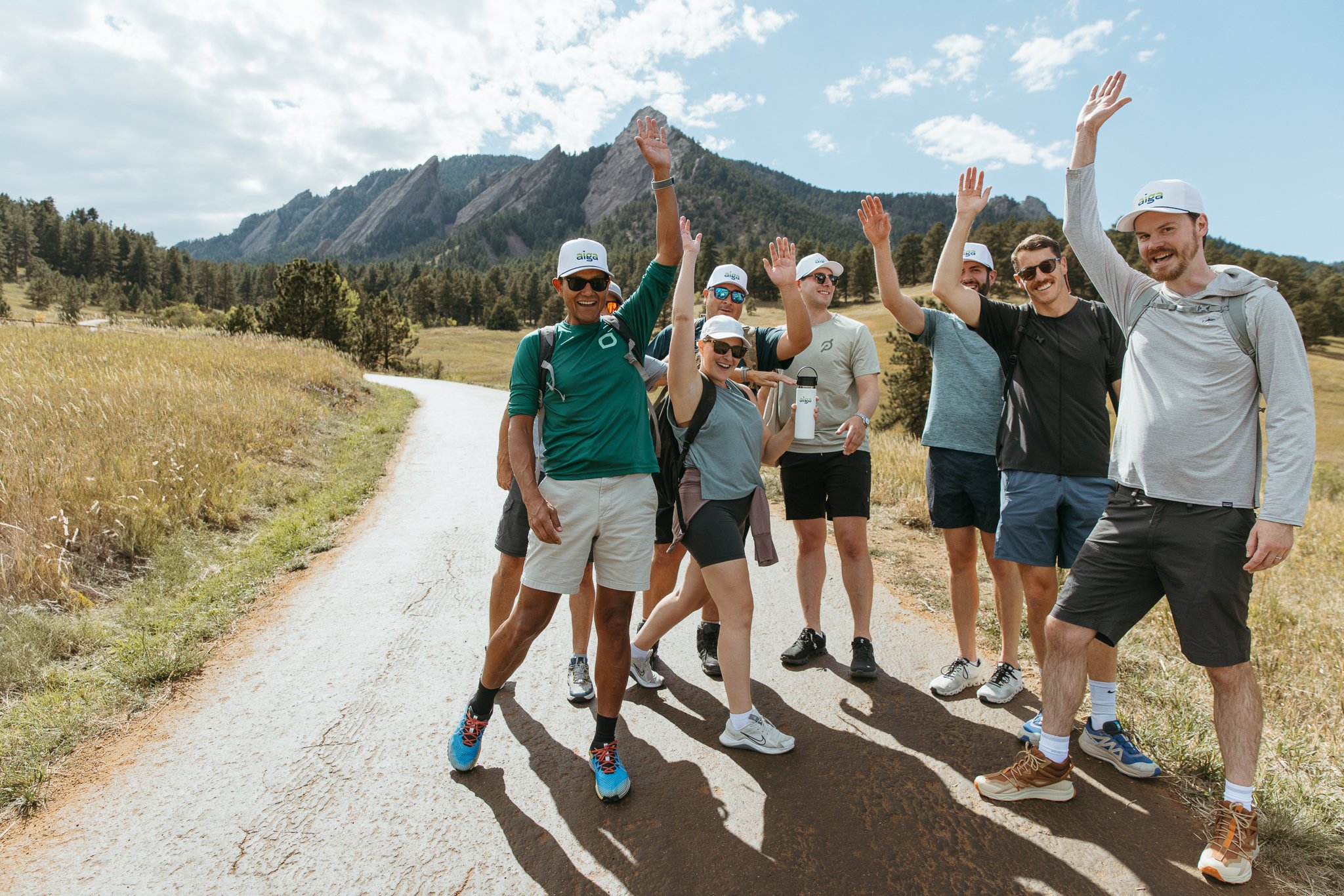 Group of people hiking on a trail in a scenic landscape with mountains, trees, and a sunny sky.