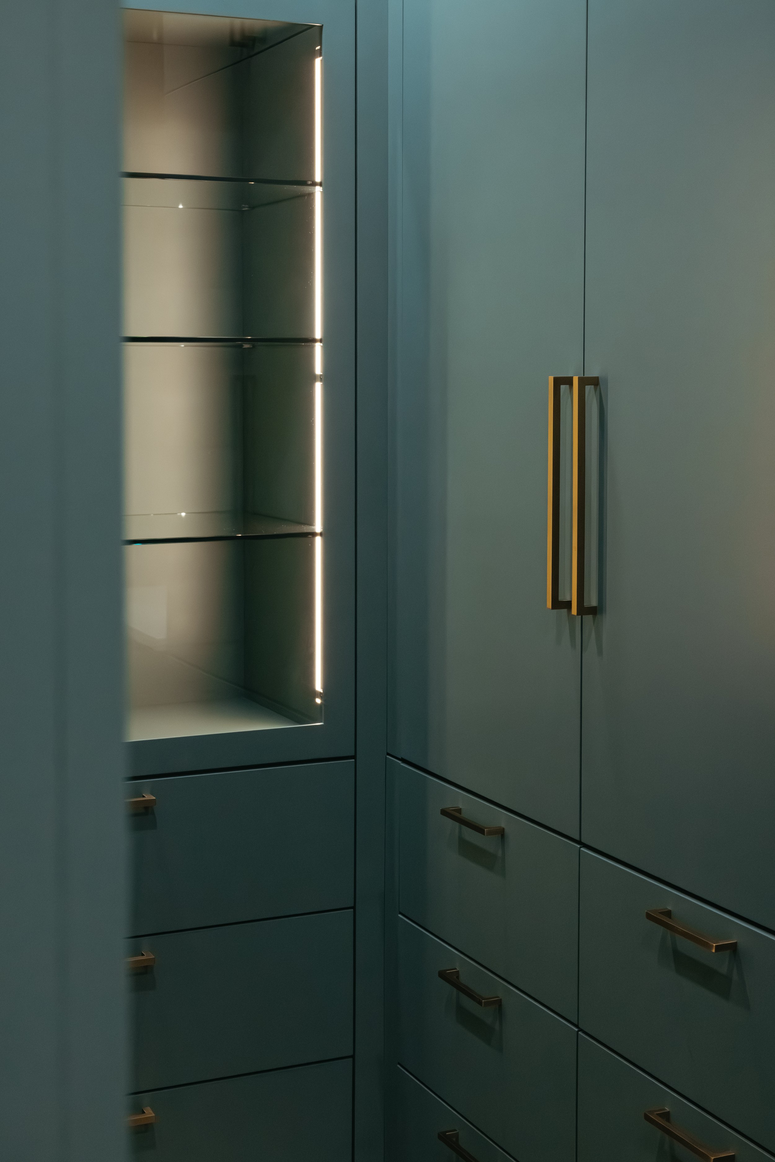 Modern minimalist closet with matte dark green cabinetry, metal handles, and glass shelves with integrated lighting.