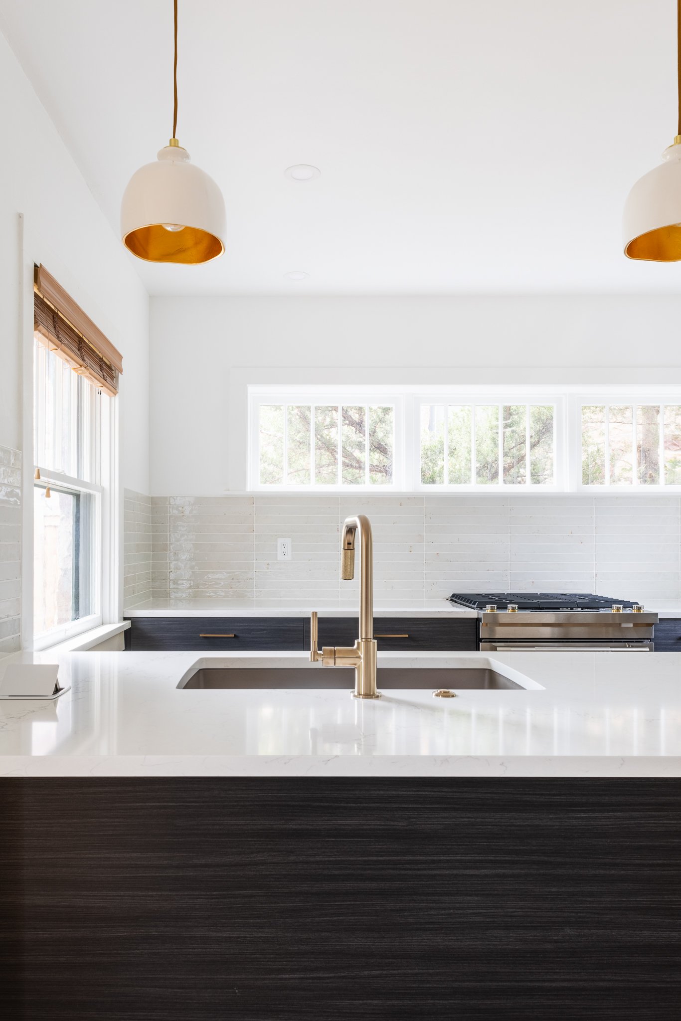 Modern kitchen interior with a gold faucet, white countertop, dark wood cabinetry, pendant lights, and large windows.