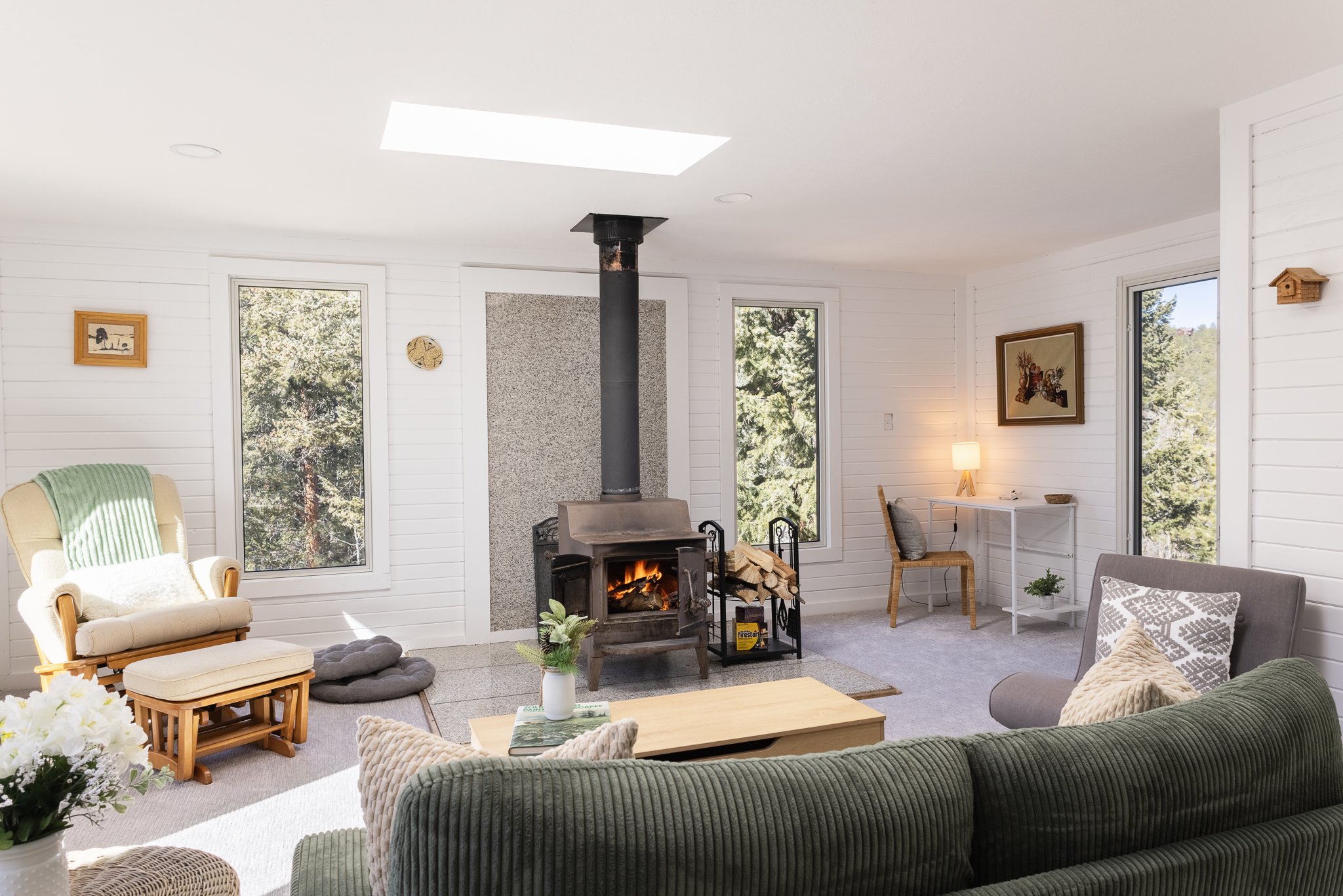 Cozy living room with a wood-burning stove, armchair, and couch, featuring large windows with forest views. A small desk with a lamp is in the corner, and decorations include framed art and plants.