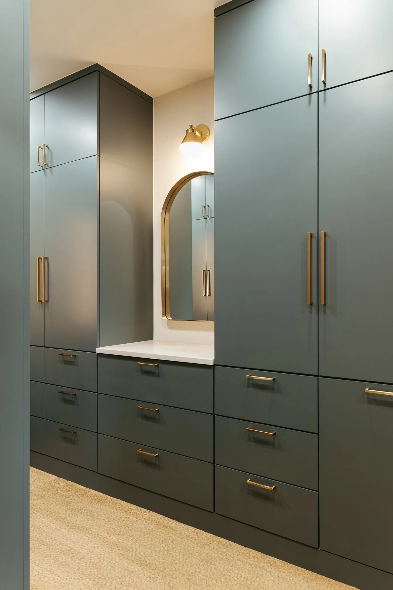 Modern closet with green cabinets, brass handles, an arched mirror, and a light fixture on textured flooring.