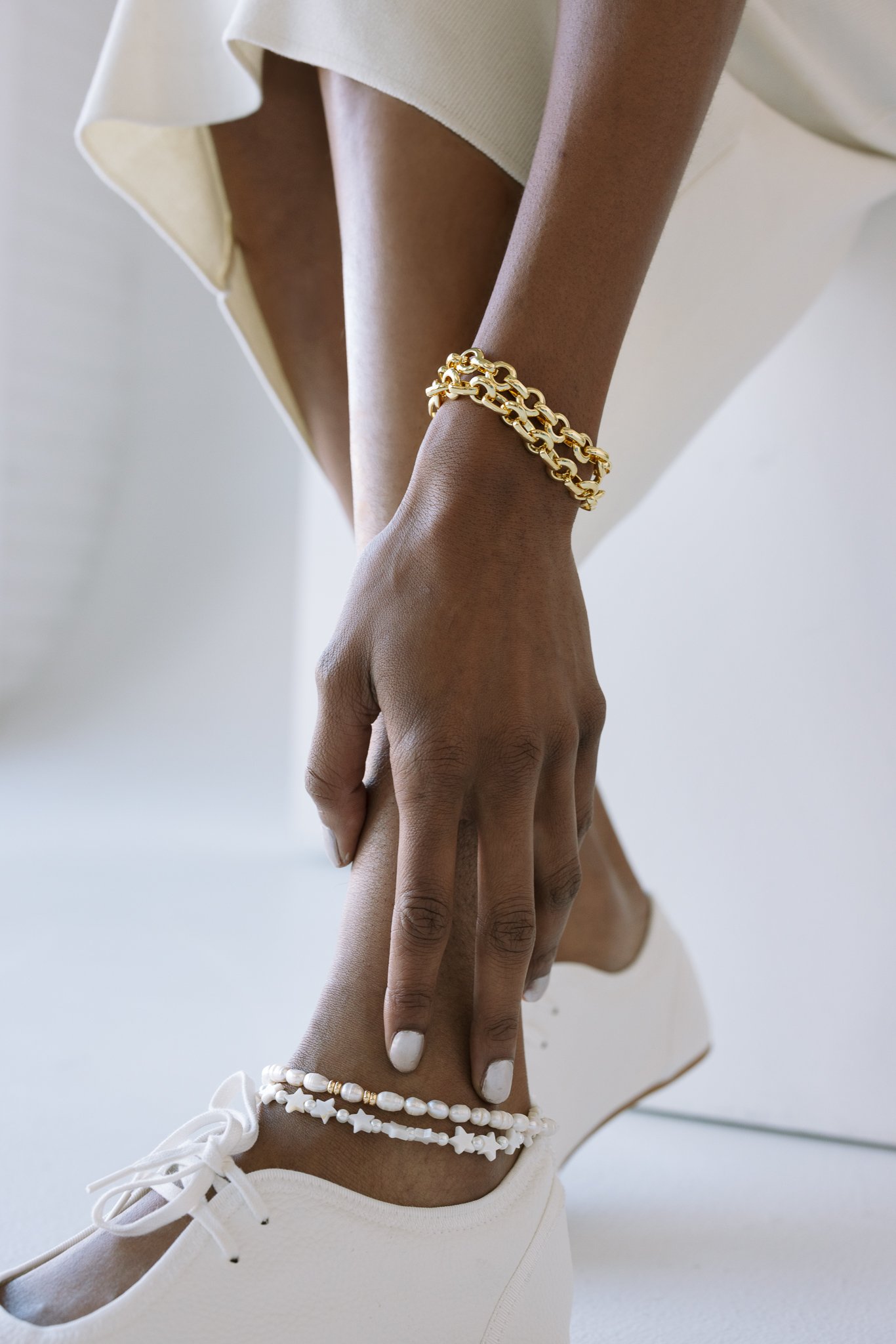 Close-up of a person's hand resting on their ankle, wearing a gold chain bracelet, a shell anklet, white shoes, and beige pants.