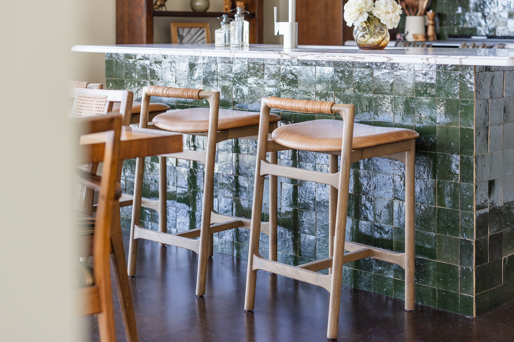 Kitchen bar area with green tiled counter, wooden barstools with leather seats, and vase with flowers on countertop.
