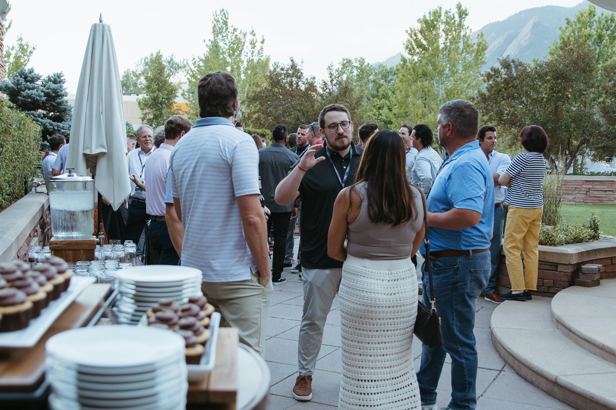 Group of people socializing outdoors at an event with mountains in the background; plates and cupcakes on a table nearby.