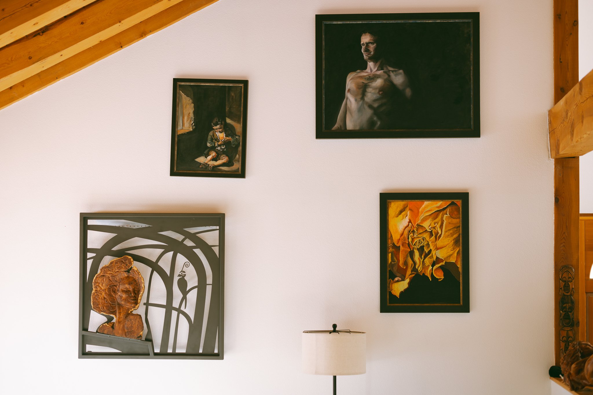 Wall with four framed artworks and a lamp. Upper artworks include a man in dark lighting and a child sitting on the floor. Lower artworks include an abstract piece with warm colors and a bronze relief of a woman's face with a bird motif.