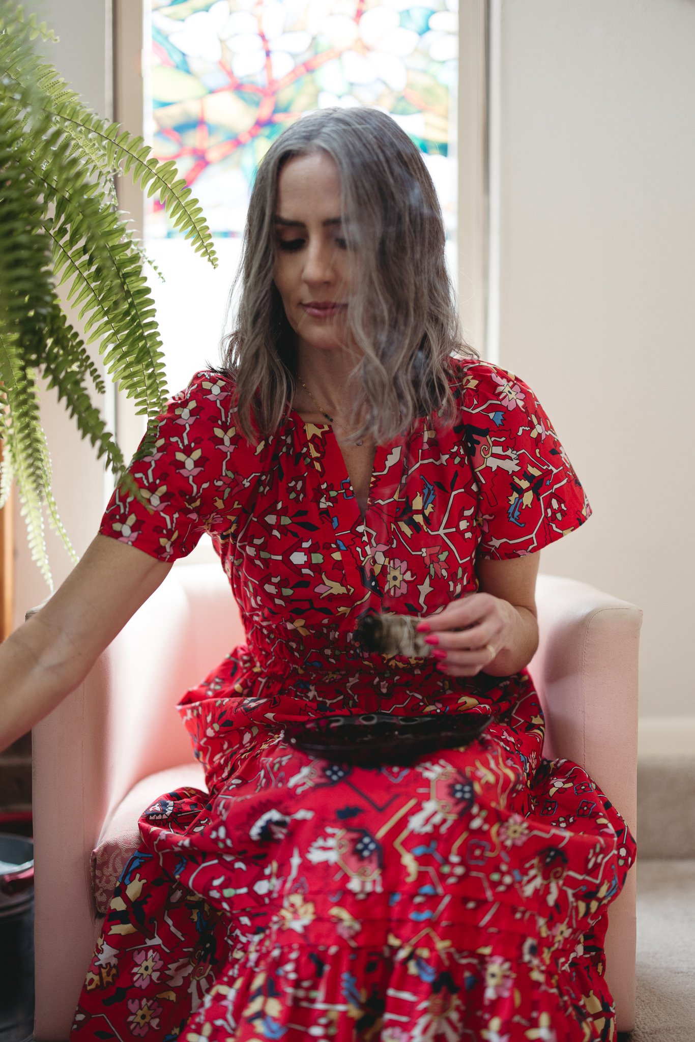 A person with gray hair wearing a vibrant red floral dress sits on a pink chair. They are holding a small plate and a smudge stick. A green fern is visible to the left, and a stained glass window is in the background.