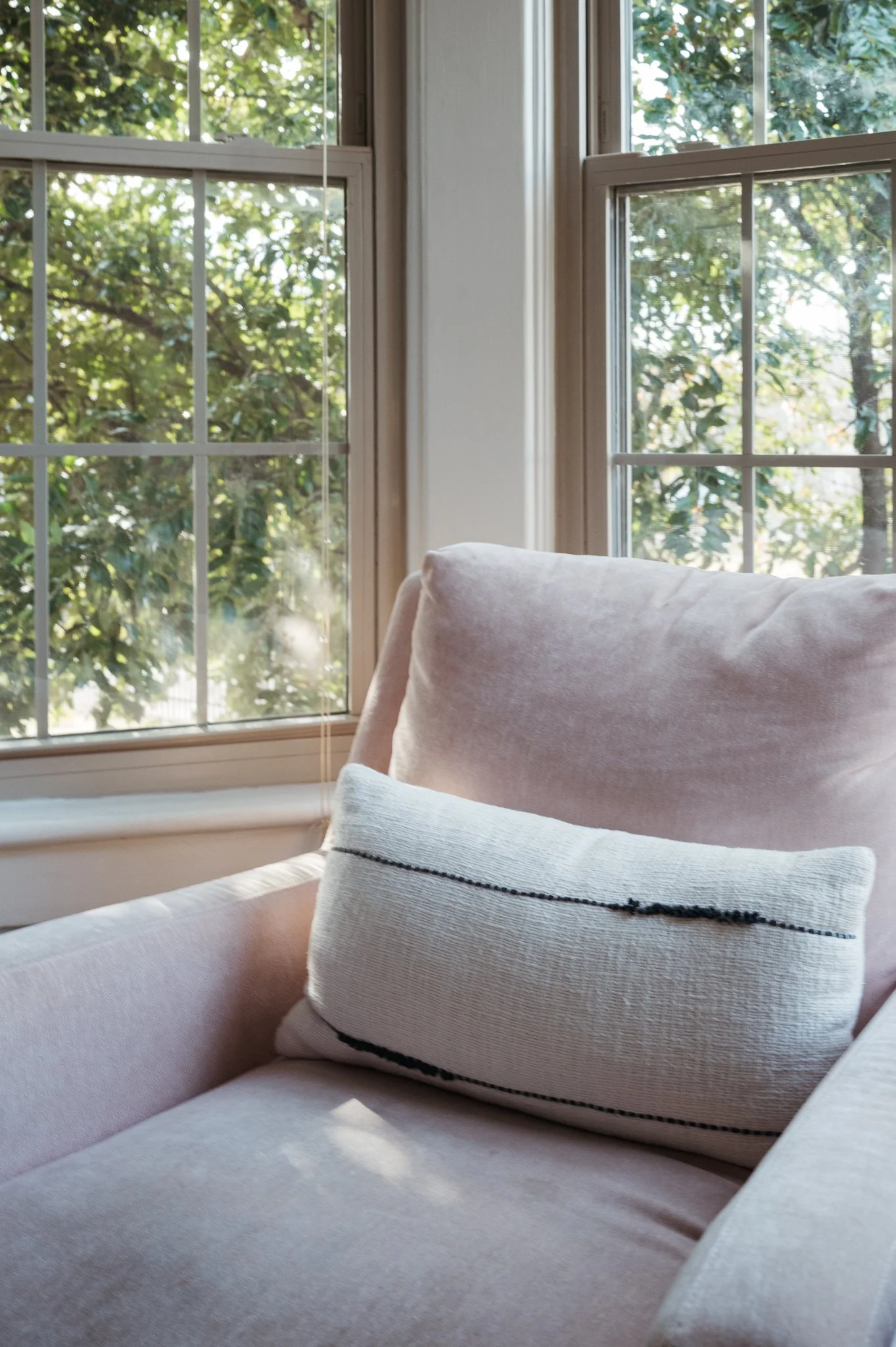 Sunlit upholstered armchair with neutral pillow beside large windows, highlighting soft natural light and a calm, inviting interior space