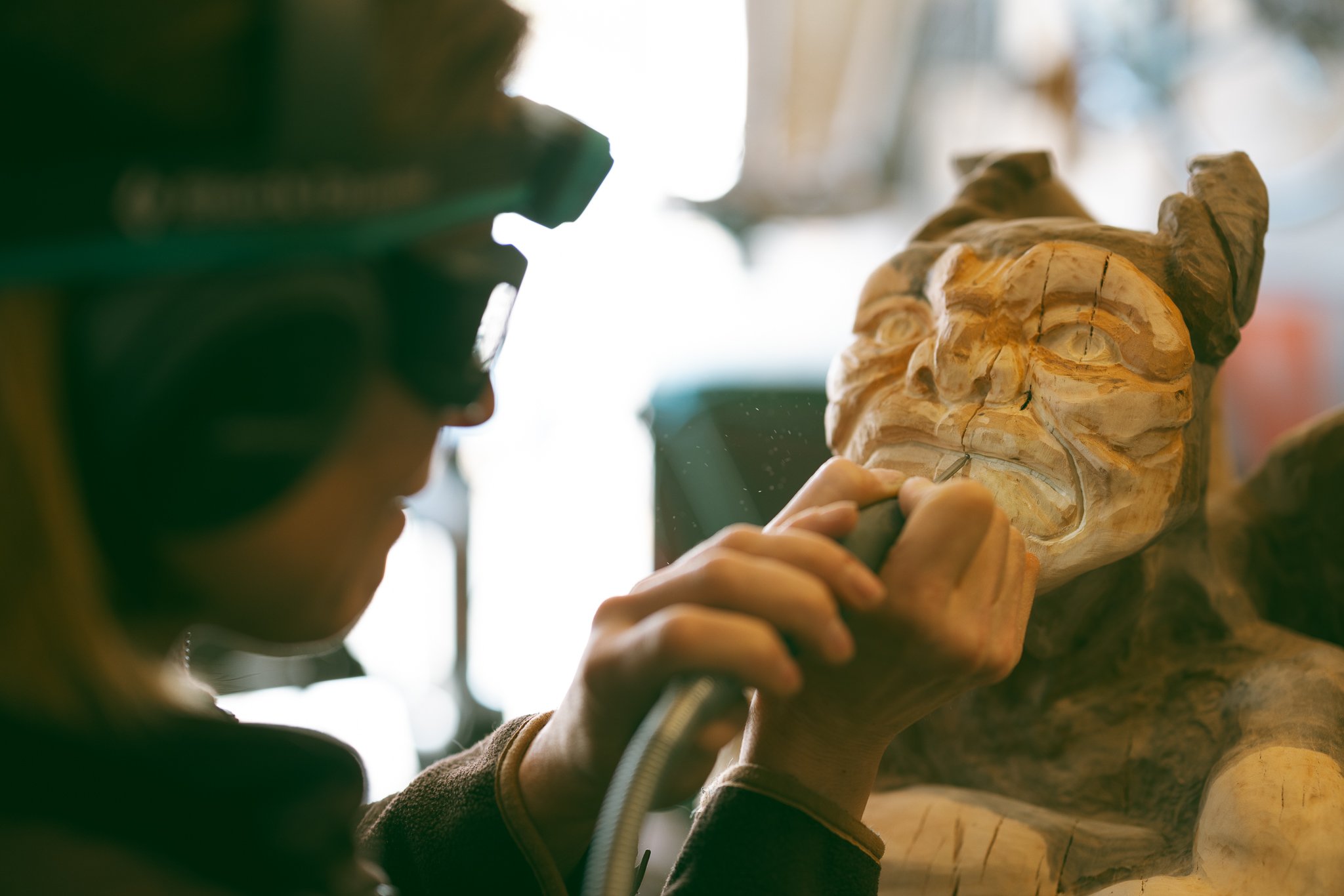 Artist carving a wooden sculpture using a power tool. The sculpture appears to be a gargoyle or mythical creature. The artist is wearing protective eyewear.