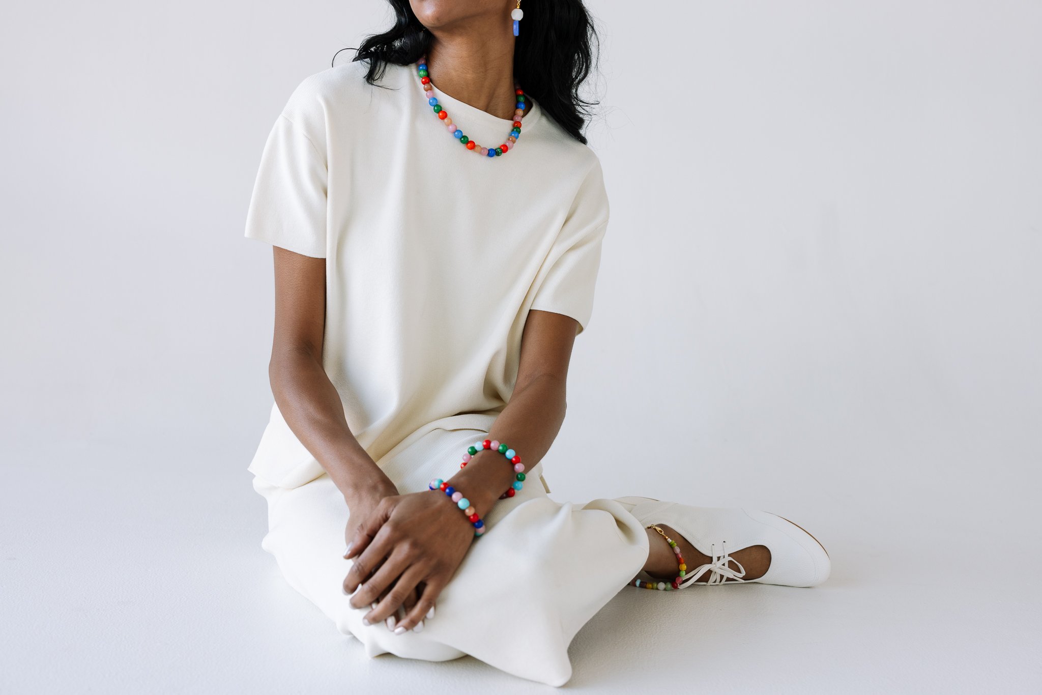 Person wearing a white outfit, colorful beaded jewelry, and white shoes on a neutral background.