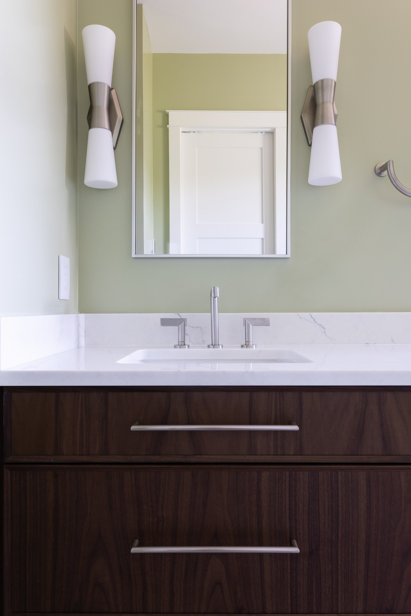 Bathroom vanity with wooden cabinet, white countertop, metal faucet, and mirror, flanked by two modern wall sconces on a light green wall.