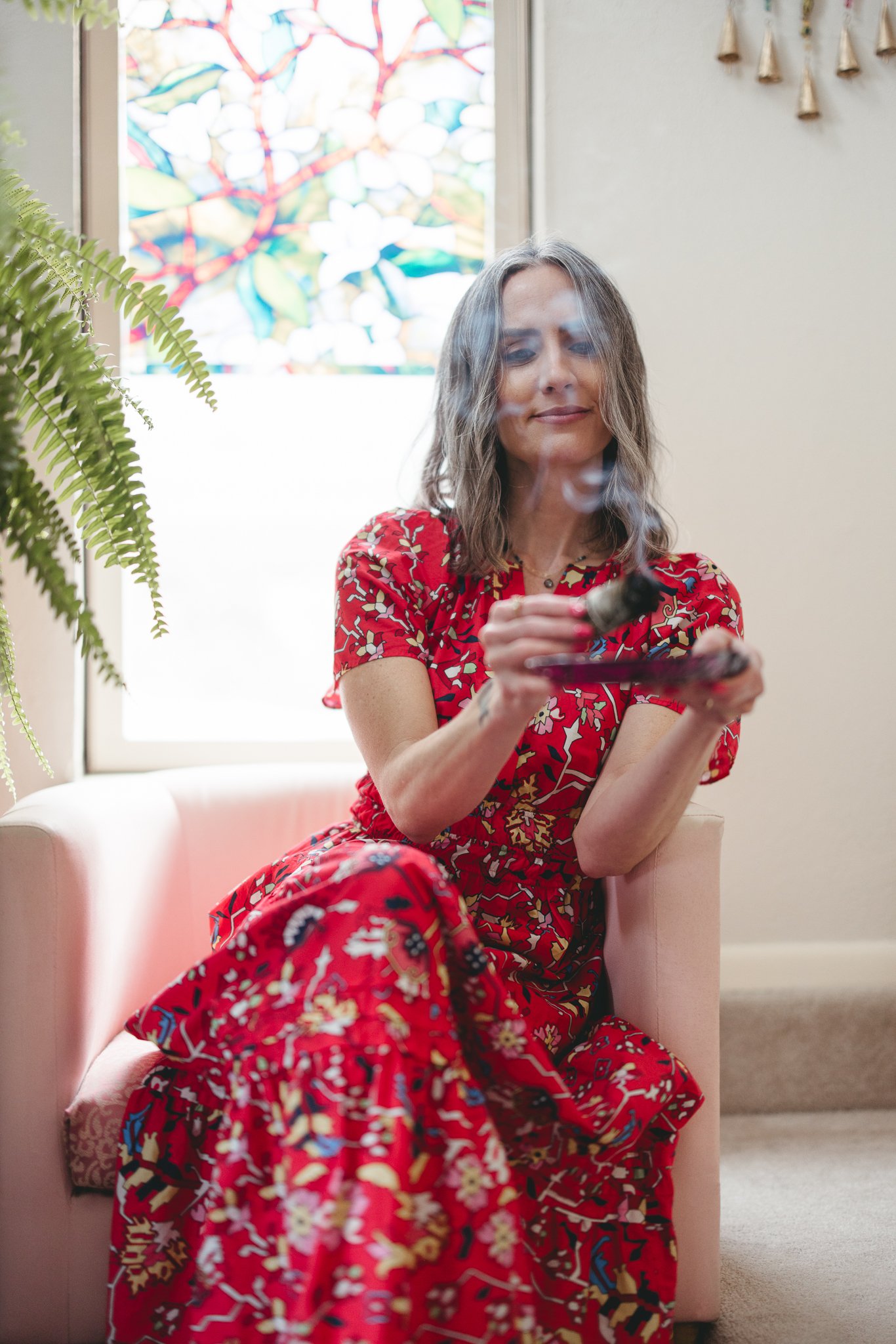 Woman in a red floral dress sitting in a chair, holding smudge stick, with a colorful stained glass window and fern in the background.