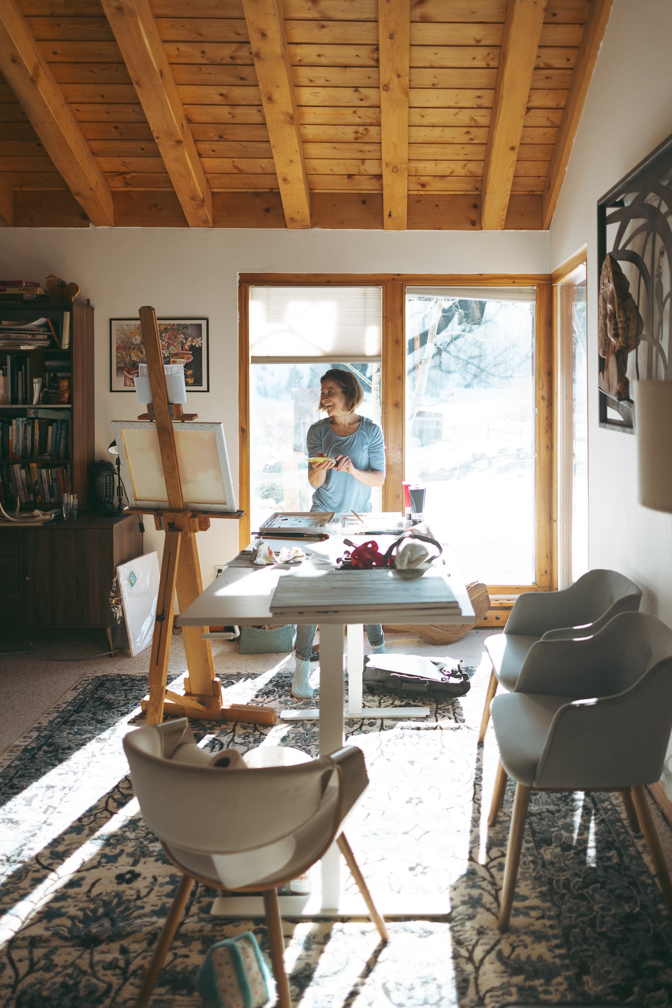 A person in a bright room with wooden ceiling beams, standing in front of a window and painting on an easel. The room contains a table, chairs, and shelves filled with books.