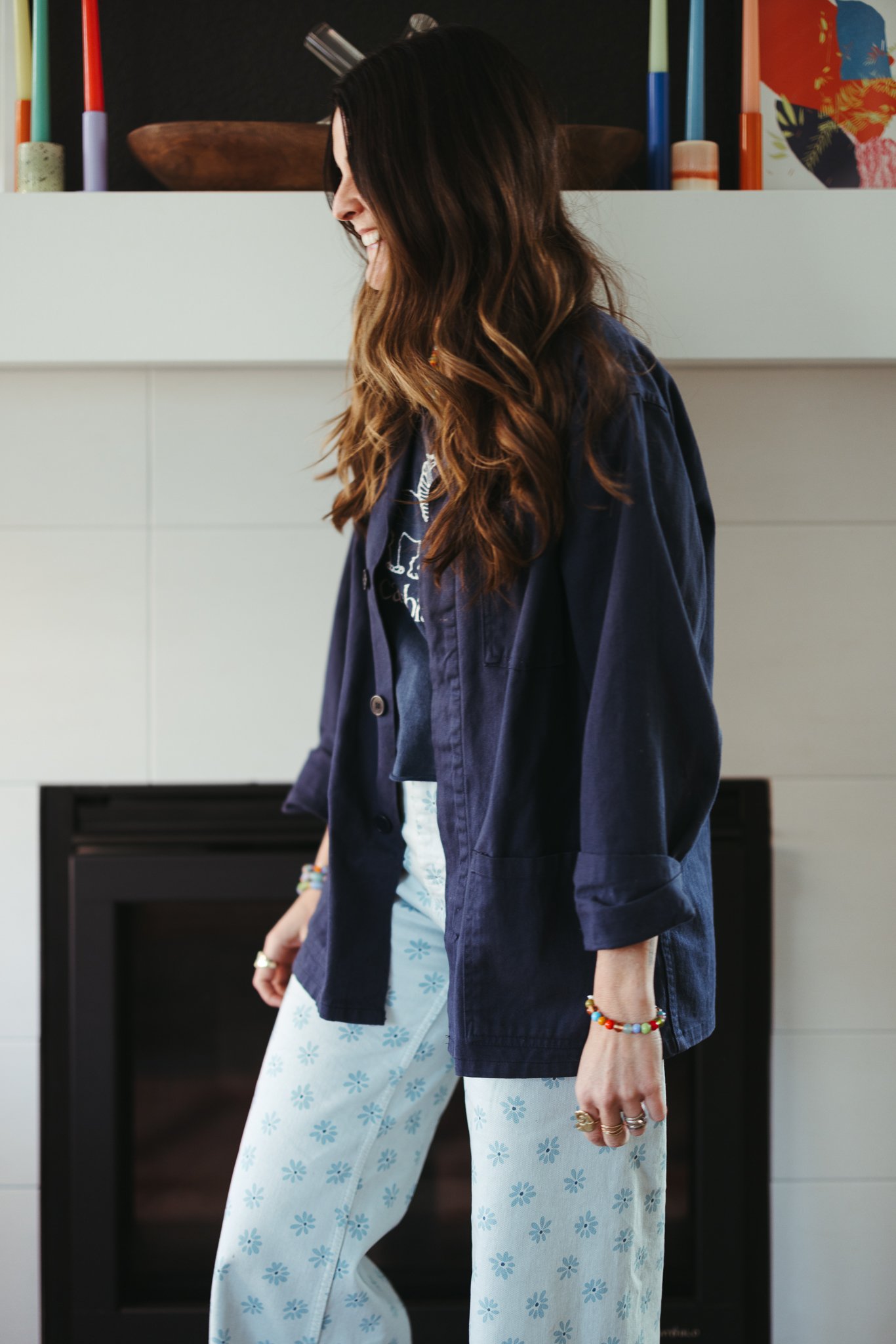 Woman wearing a navy jacket over a graphic T-shirt and floral patterned pants, standing near a white mantel with colorful candles.