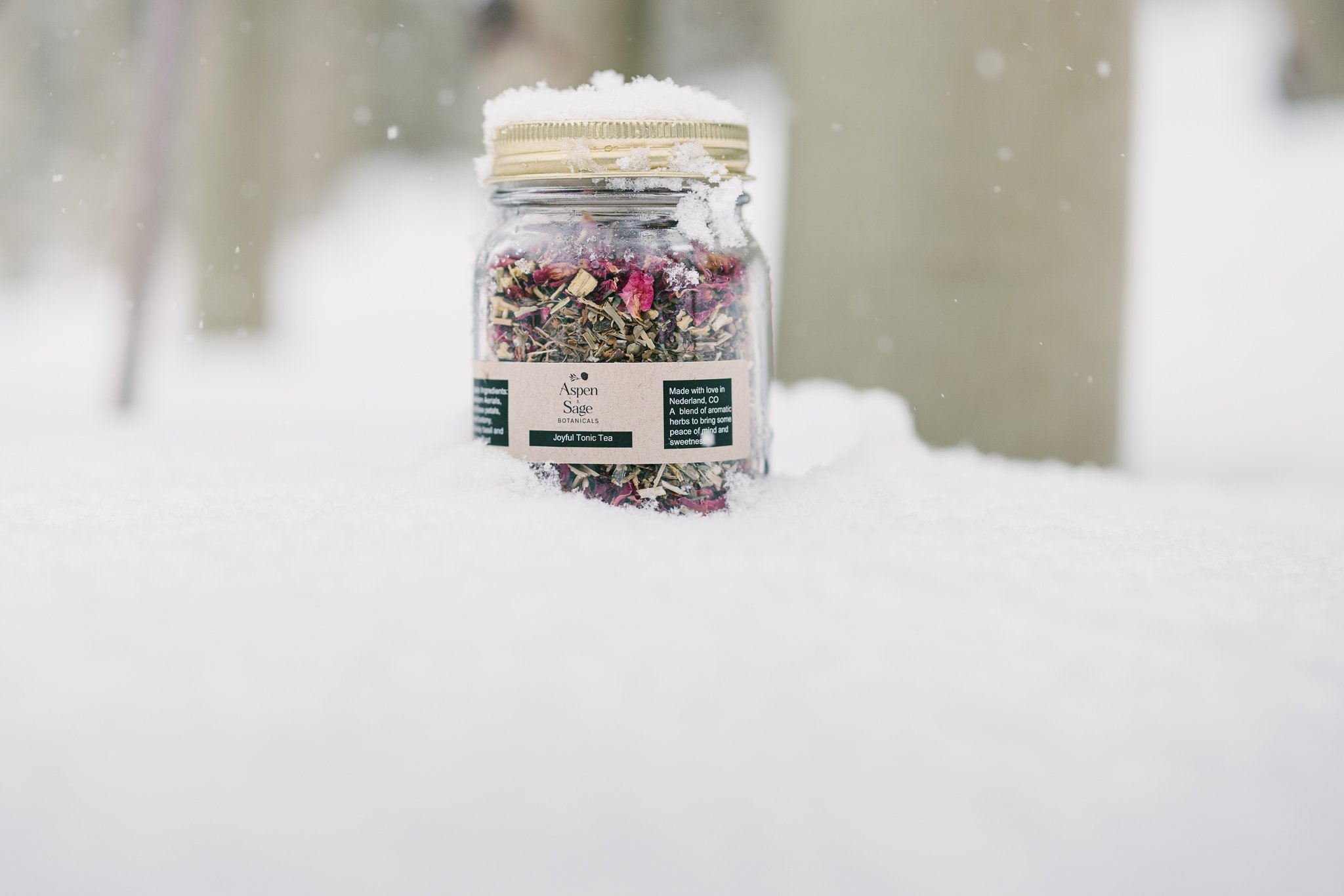 A jar of Aspen Sage Botanicals Joyful Tonic Tea sitting in the snow, with a gold lid and labeled with ingredients and product information. The background is snowy and out of focus.