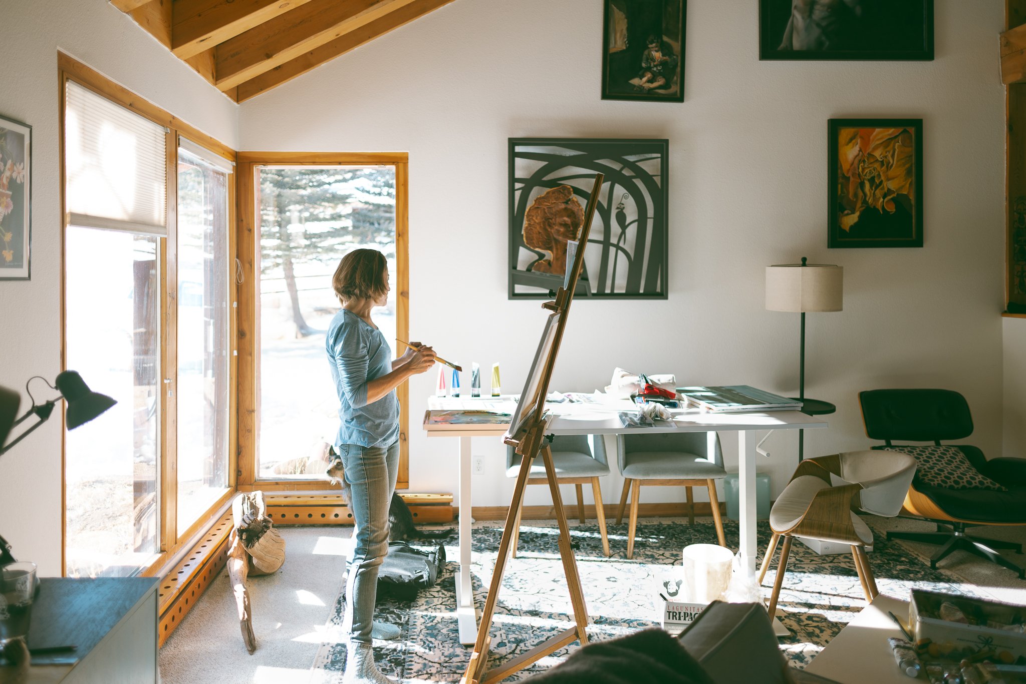 Person painting on a canvas in a bright room with wooden ceiling and large windows, surrounded by art supplies and furniture, with paintings on the walls.