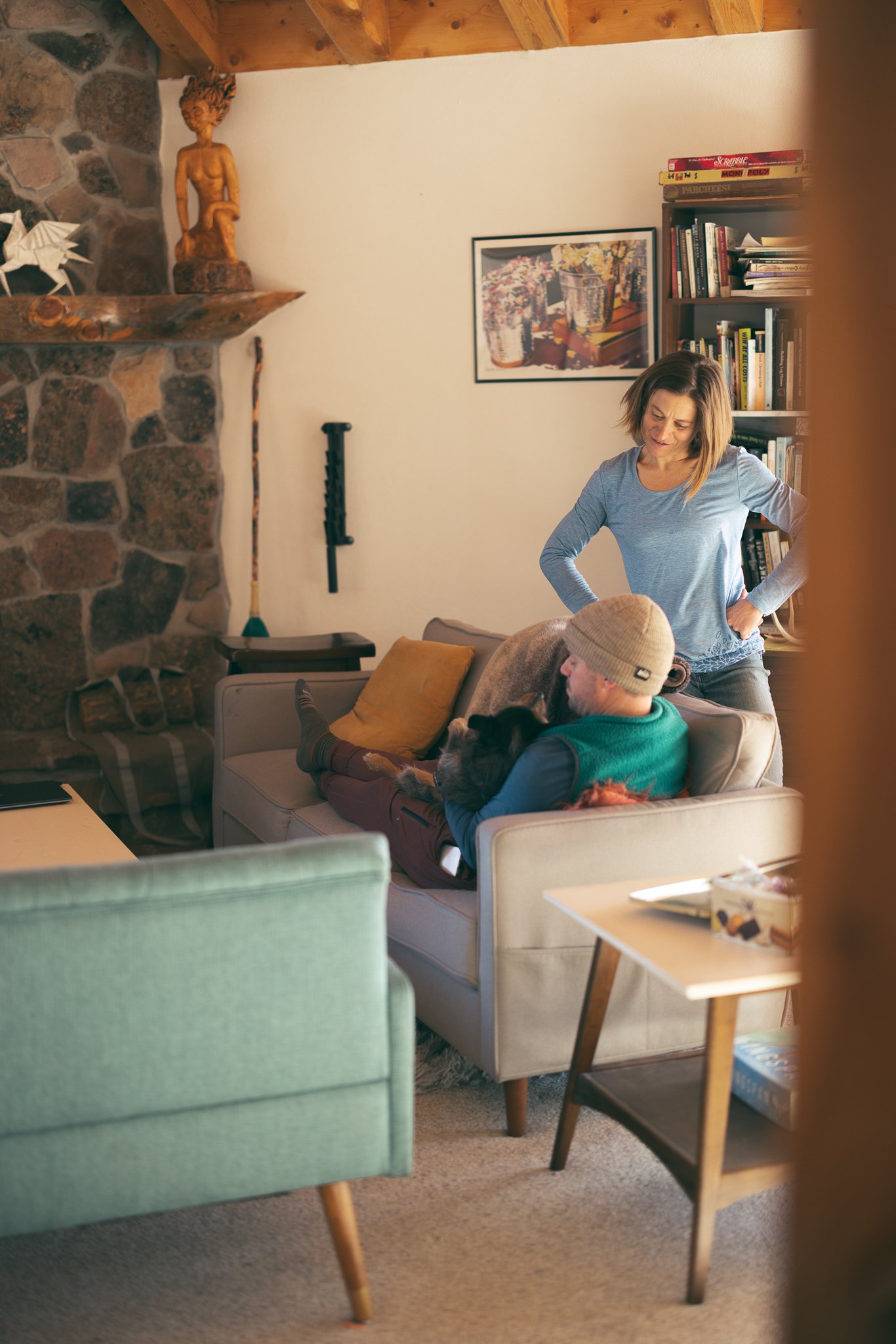 Cozy living room with a stone fireplace, two people interacting, one seated on a sofa with a dog, and the other standing. Bookshelves and a side table are visible, with eclectic decorations.