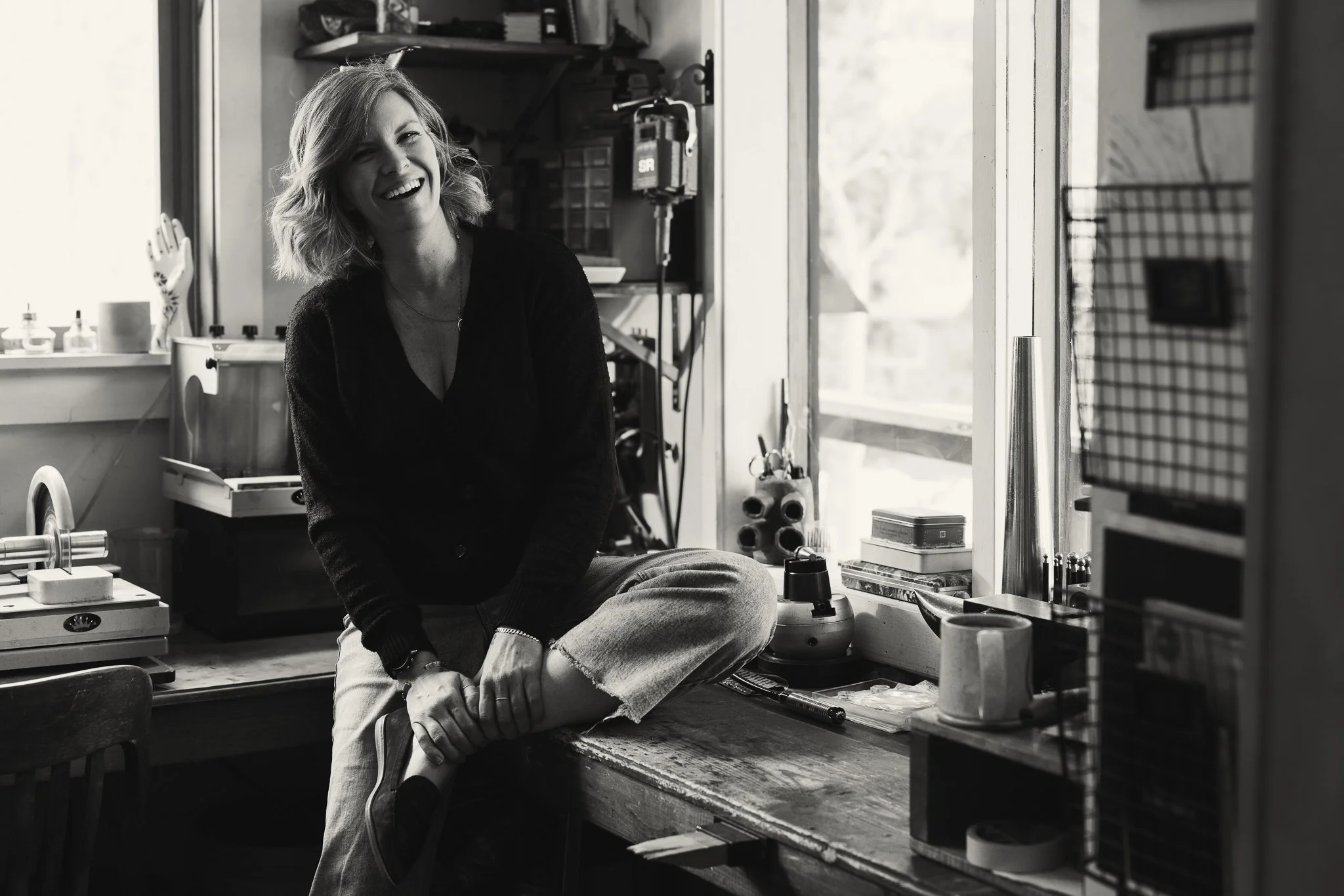 Black and white photo of a person sitting on a workbench in a workshop, surrounded by tools and equipment, with a window in the background.