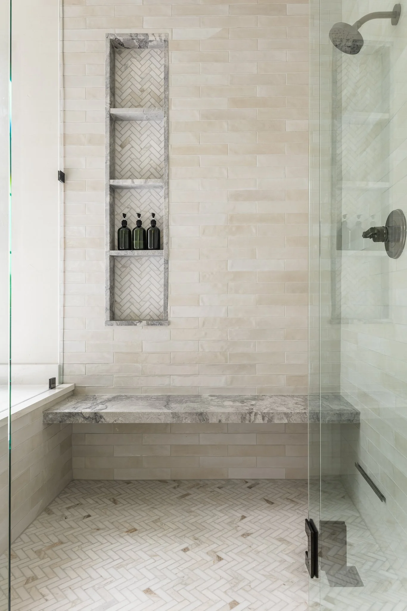 Modern shower with beige subway tile walls, marble bench, herringbone tile floor, and built-in shelves holding three black bottles.
