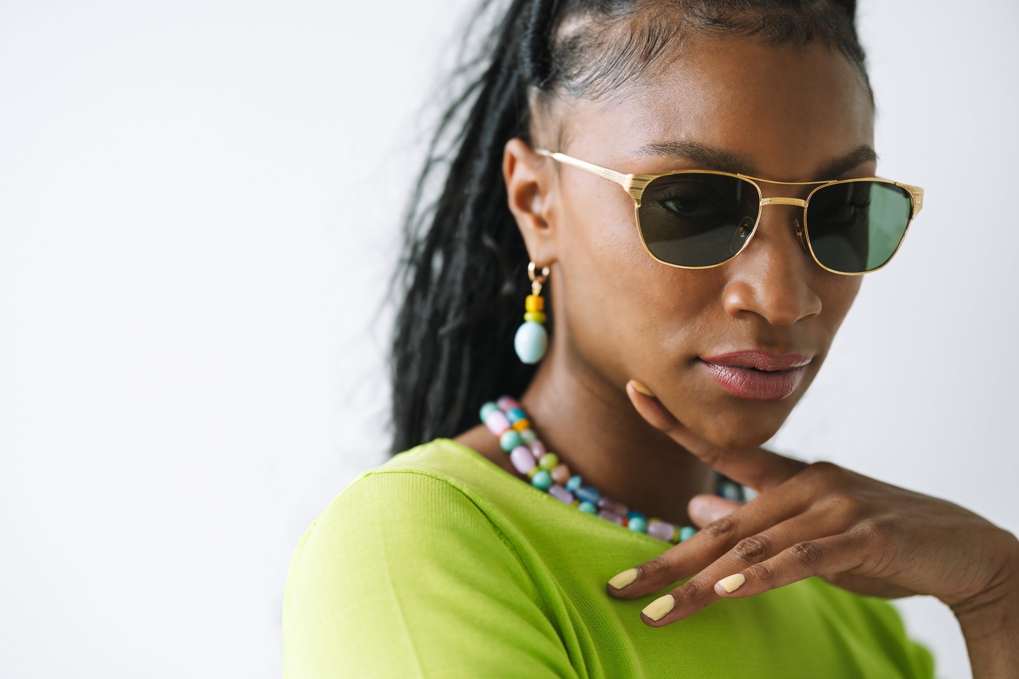 Woman in sunglasses wearing a green shirt, colorful beaded necklace, and earrings, posing with her hand near her chin.